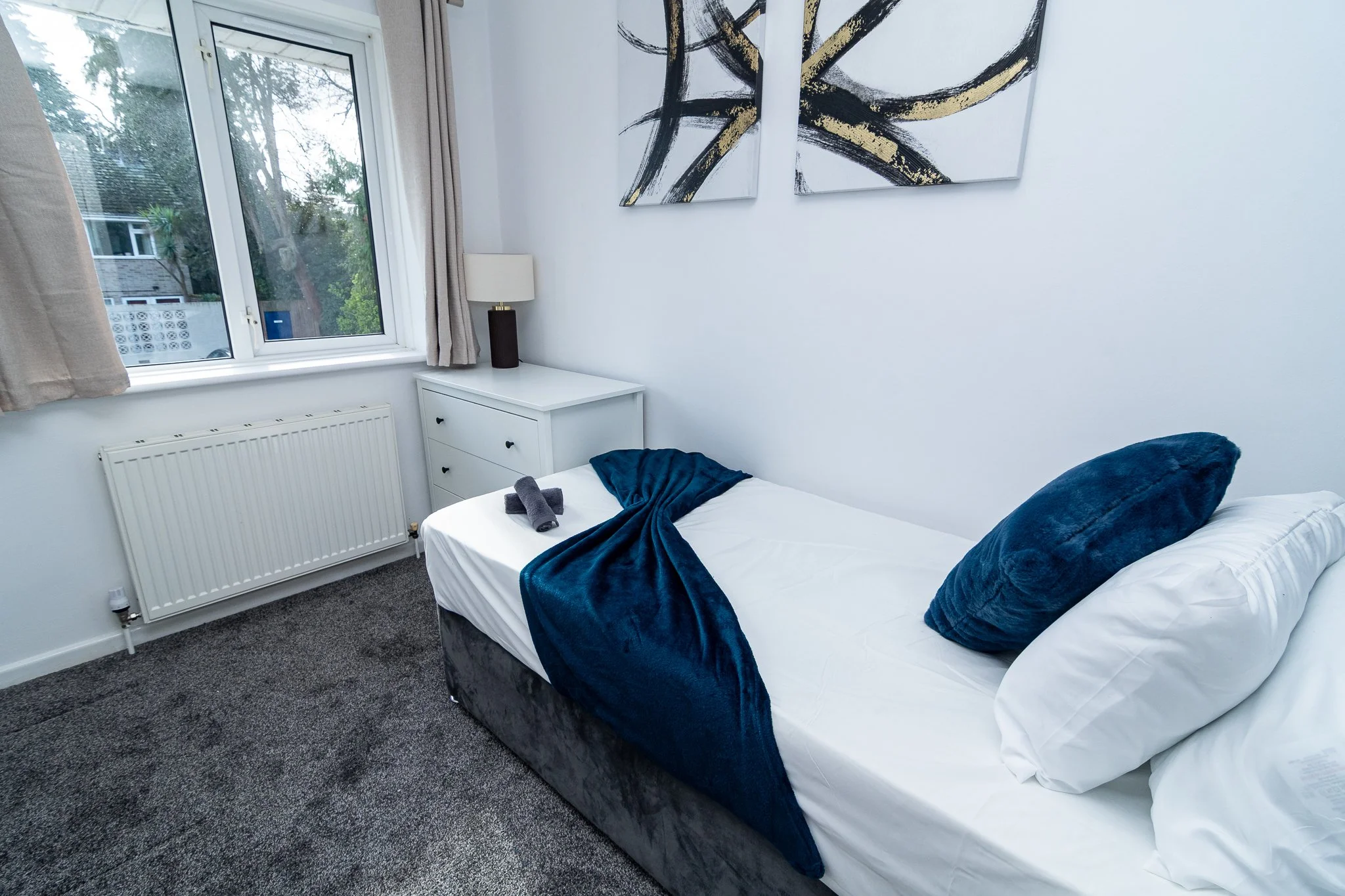 A bedroom featuring a bed with white linens and navy blue accents, a window with beige curtains, a white dresser with a lamp, and abstract art on the wall.