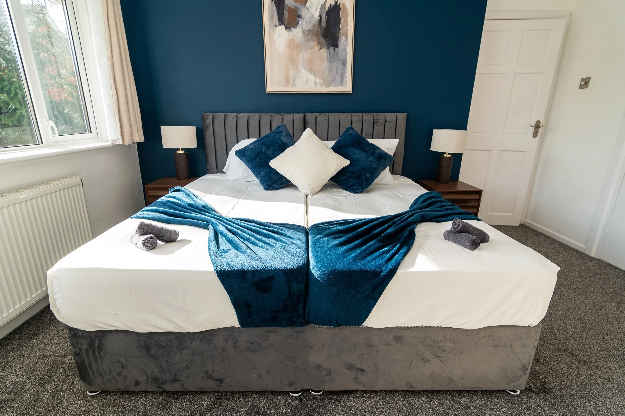 A neatly made bed with a velvet gray headboard, topped with decorative pillows in shades of blue and beige, and a dark blue throw blanket folded at the foot. There are two bedside tables with white lamps, a window with white curtains, and a large abstract painting on the blue accent wall behind the bed.