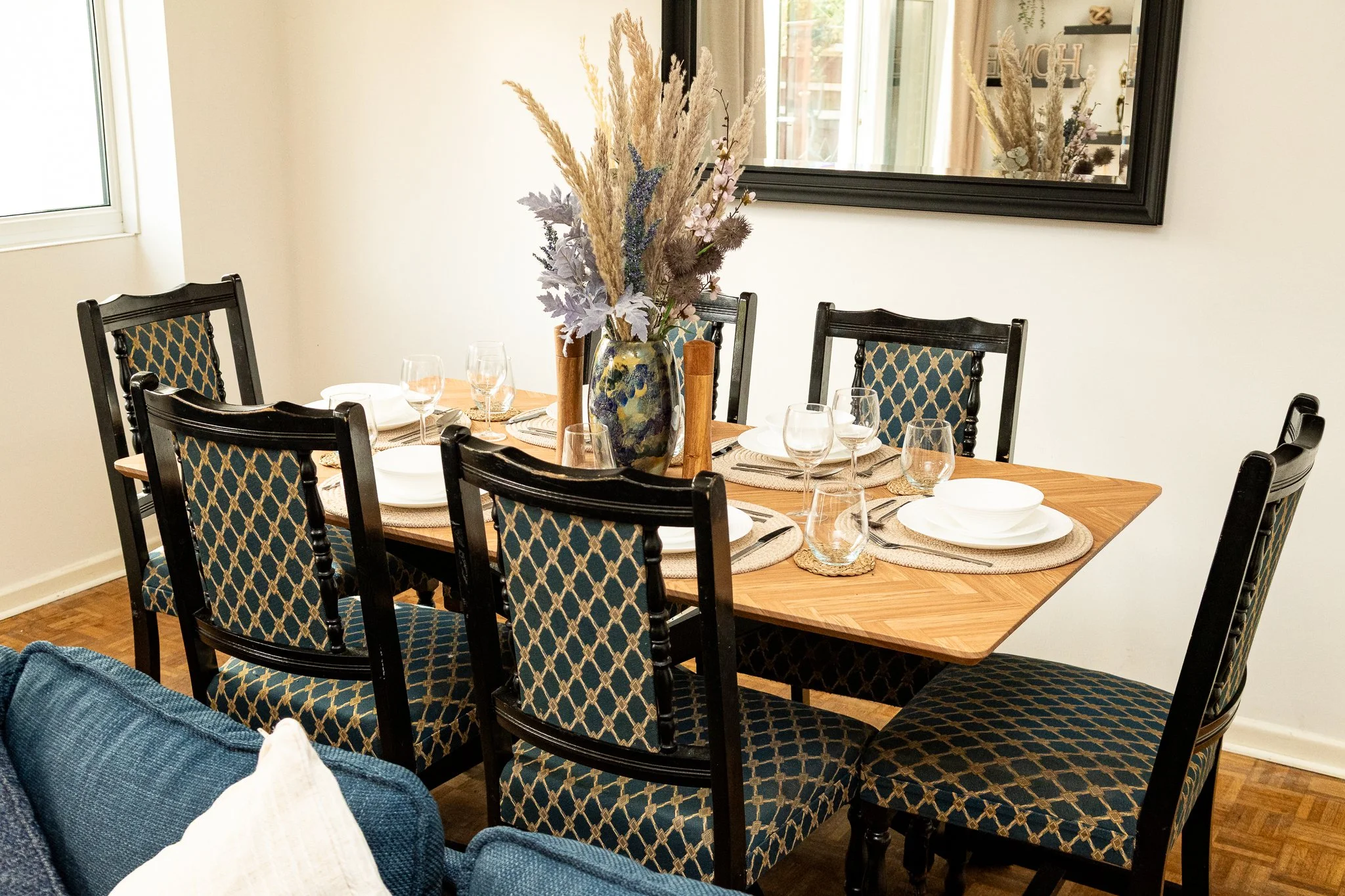 A dining room table set with plates, glasses, and placemats, featuring a tall vase with dried flowers as a centerpiece. The room has a window, a large mirror on the wall, and wooden chairs with patterned upholstery around the table.
