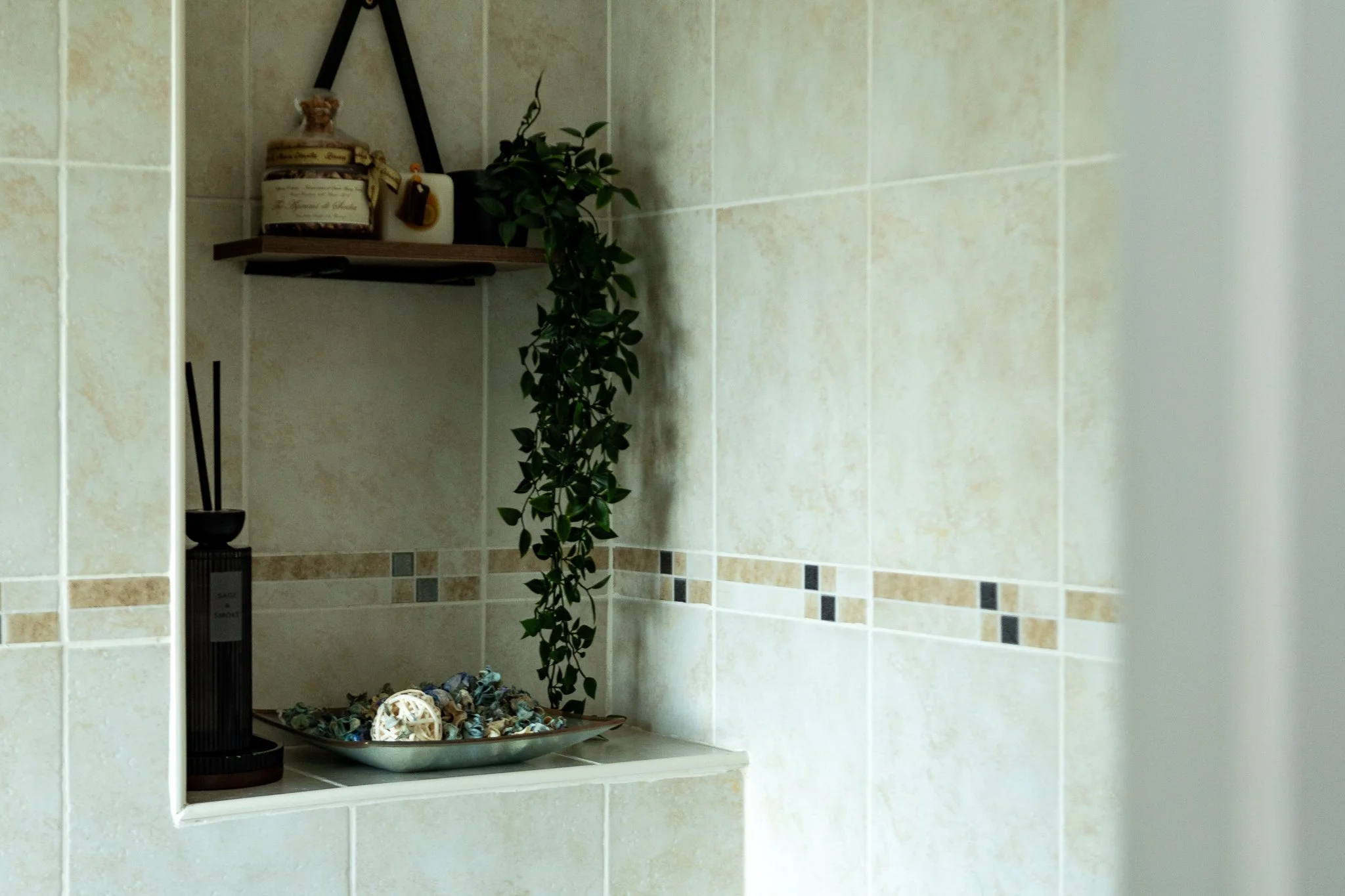 Bathroom shower niche with beige tiles, a hanging green plant, a decorative jar, a white drawer, a black diffuser, and a dish filled with colorful stones and decorative objects.