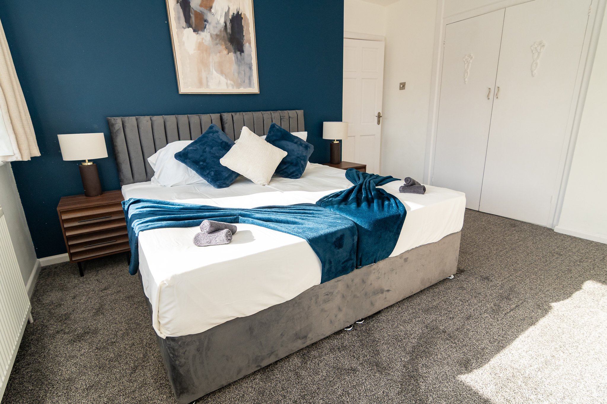 A bedroom with a bed that has a gray upholstered headboard, blue and white pillows, a blue throw blanket, and neatly folded towels. There are two matching bedside tables with white lamps. The wall behind the bed is painted dark blue, with a large abstract painting hanging above. To the right, there's a white closet with decorative handles and a closed white door. The floor is carpeted in gray.