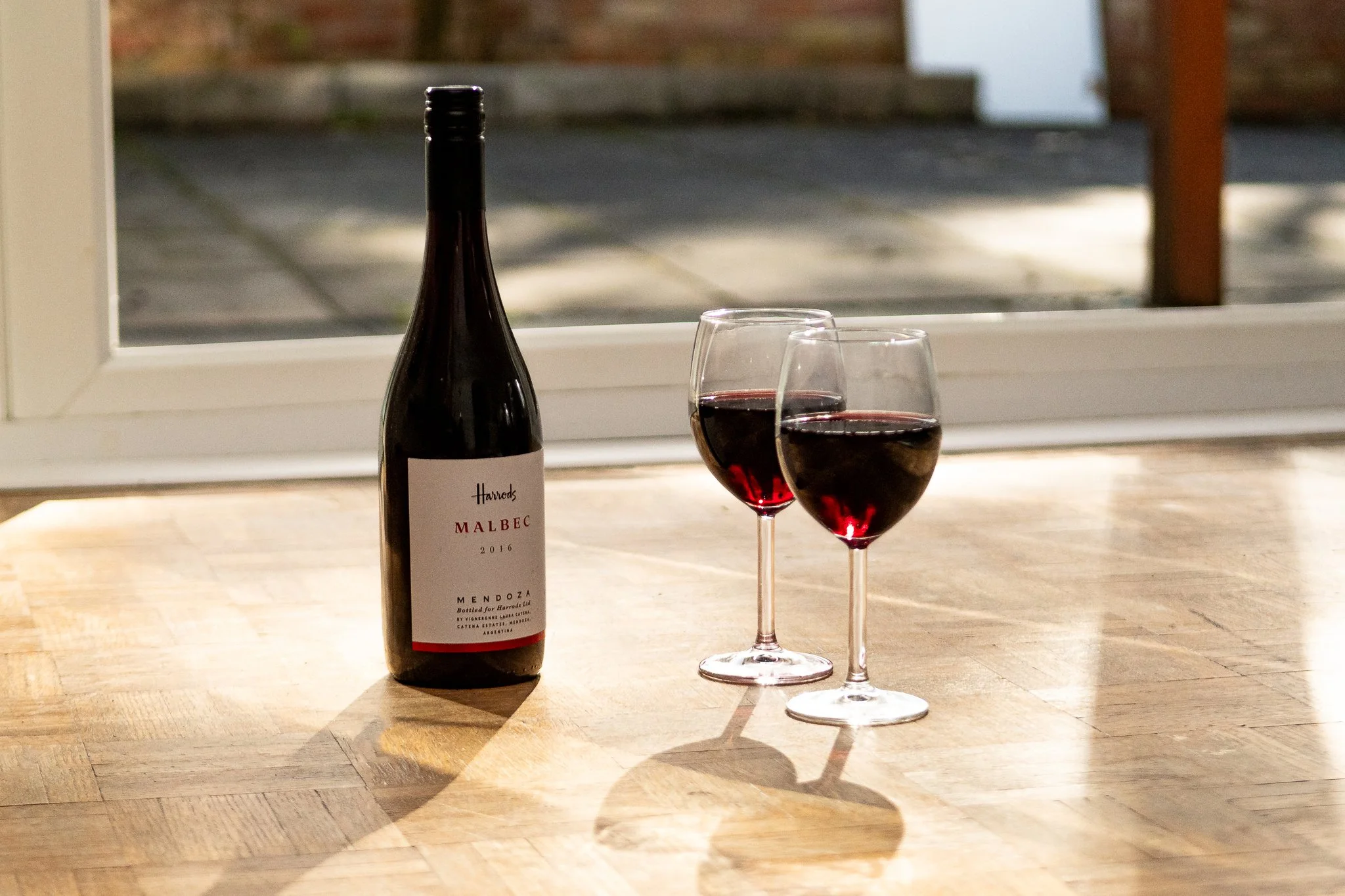 A bottle of red Malbec wine and two glasses filled with red wine on a wooden surface in front of a window with a blurry outdoor scene.