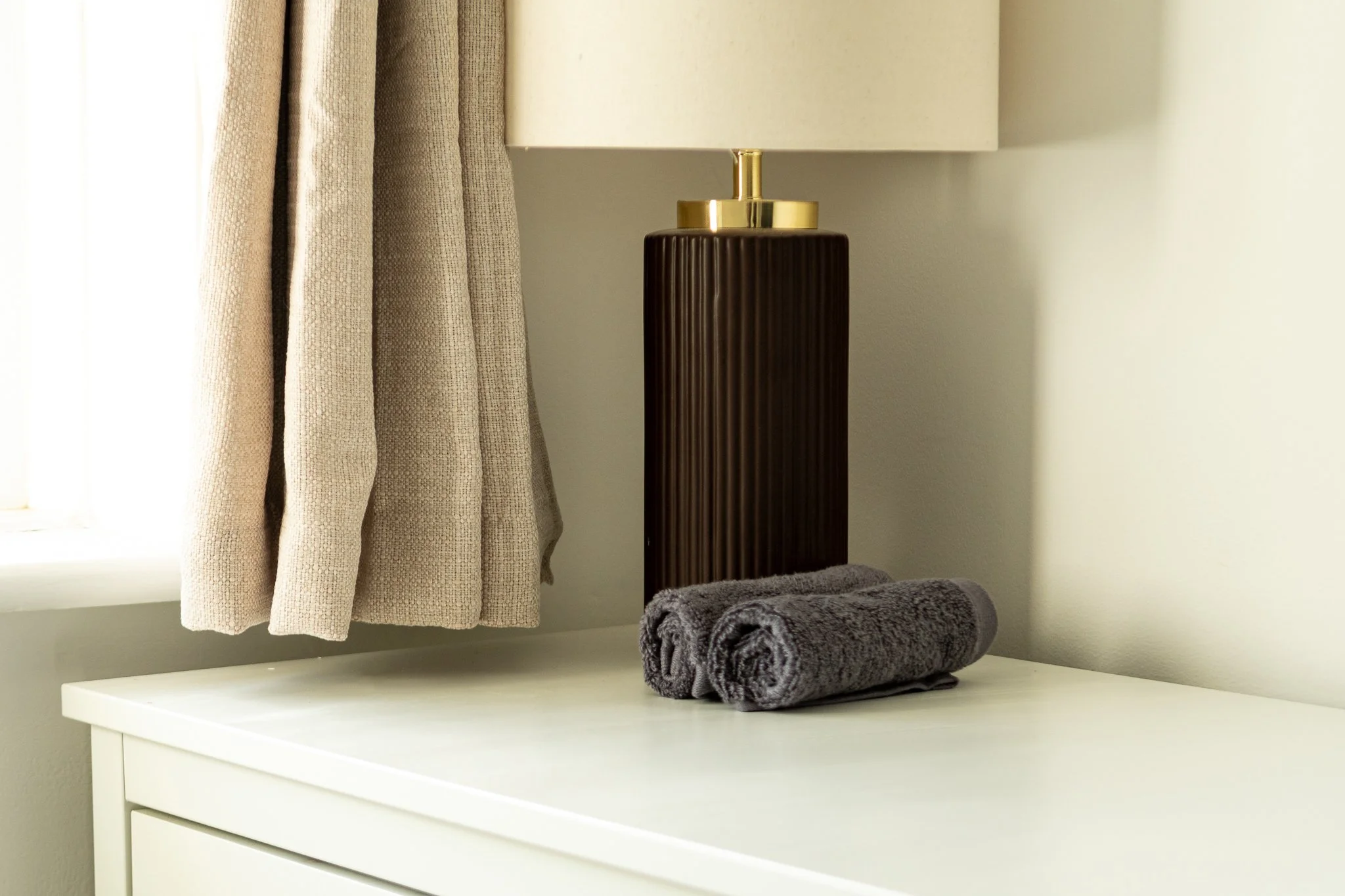 A white dresser with three drawers, a rolled dark gray towel on top, a table lamp with a white shade and gold accent, beige curtains, and a window in the background.