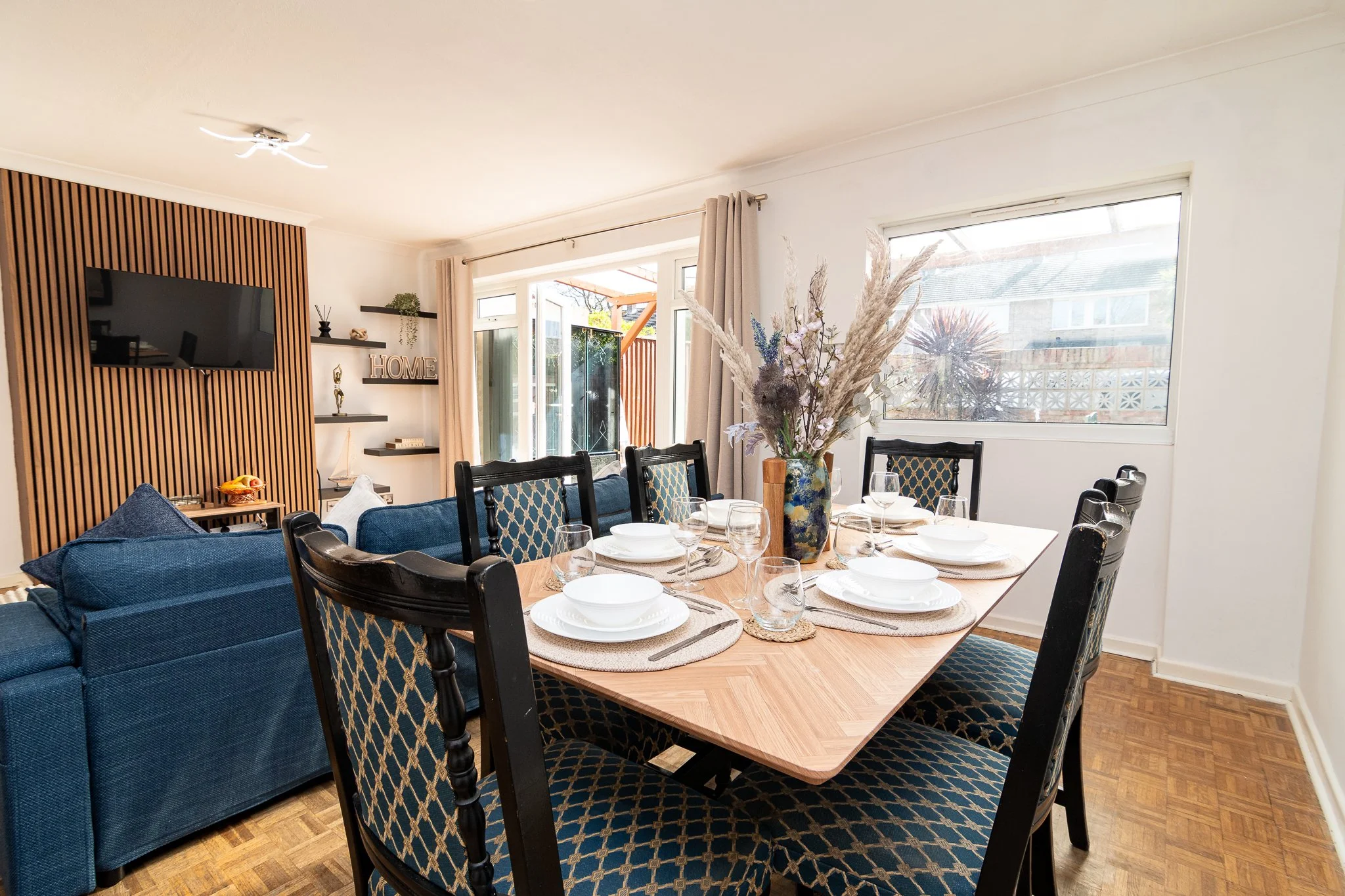 A dining area with a natural wood table set with white dishes, glasses, and woven placemats. Surrounding the table are black and blue patterned chairs. The room has large windows with beige curtains, a vase with dried flowers, and a living space with a blue couch and wall-mounted TV on a wood-paneled accent wall.