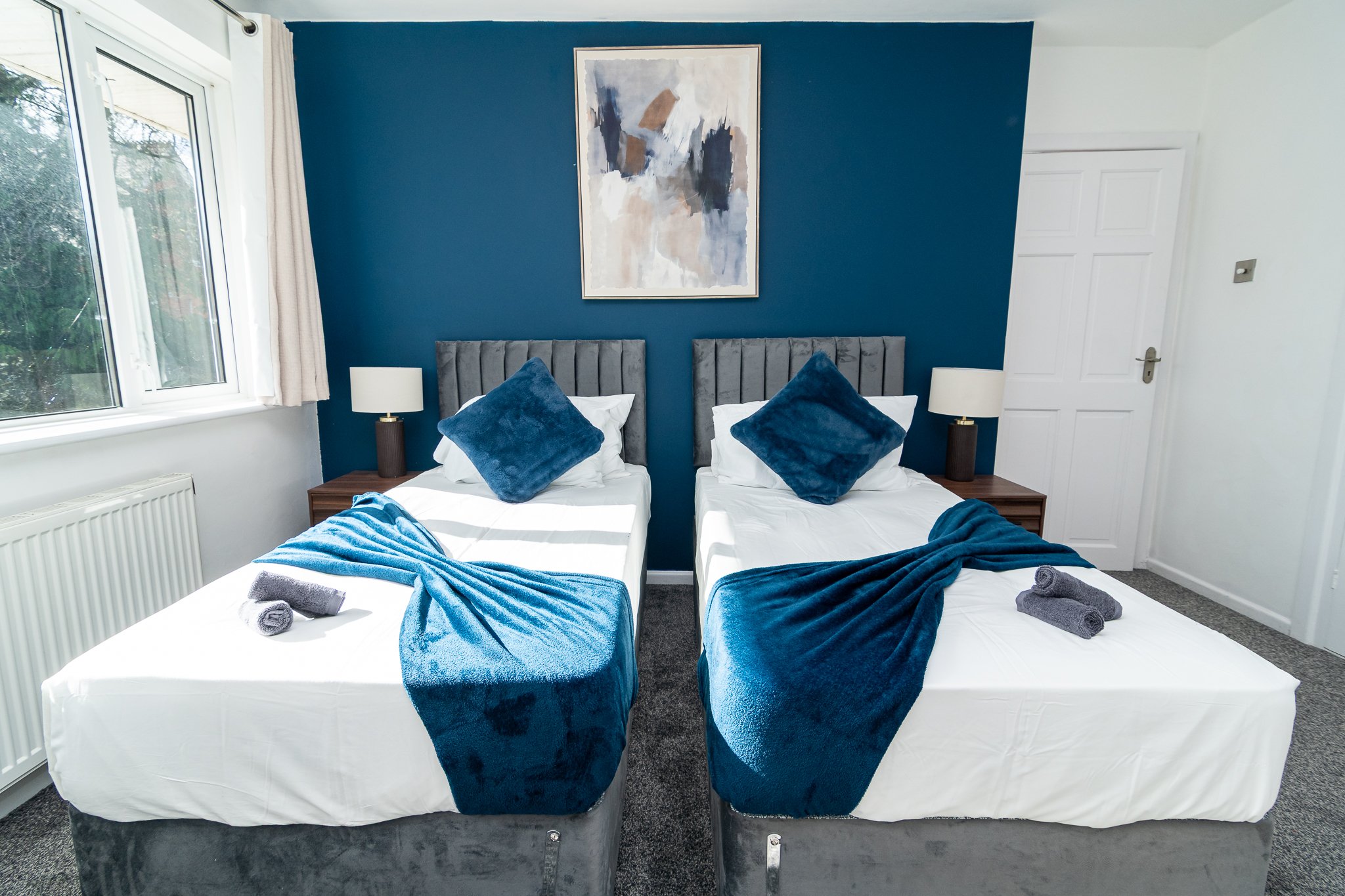 A bedroom with two single beds, each with white sheets, dark gray bed skirts, and blue throws at the foot. The beds have matching gray upholstered headboards and large blue pillows. A wooden nightstand with a white lamp is placed between the beds. A large abstract painting with shades of blue, white, black, and beige hangs above the beds. A window with white curtains is on the left, and a white door is on the right side of the room.