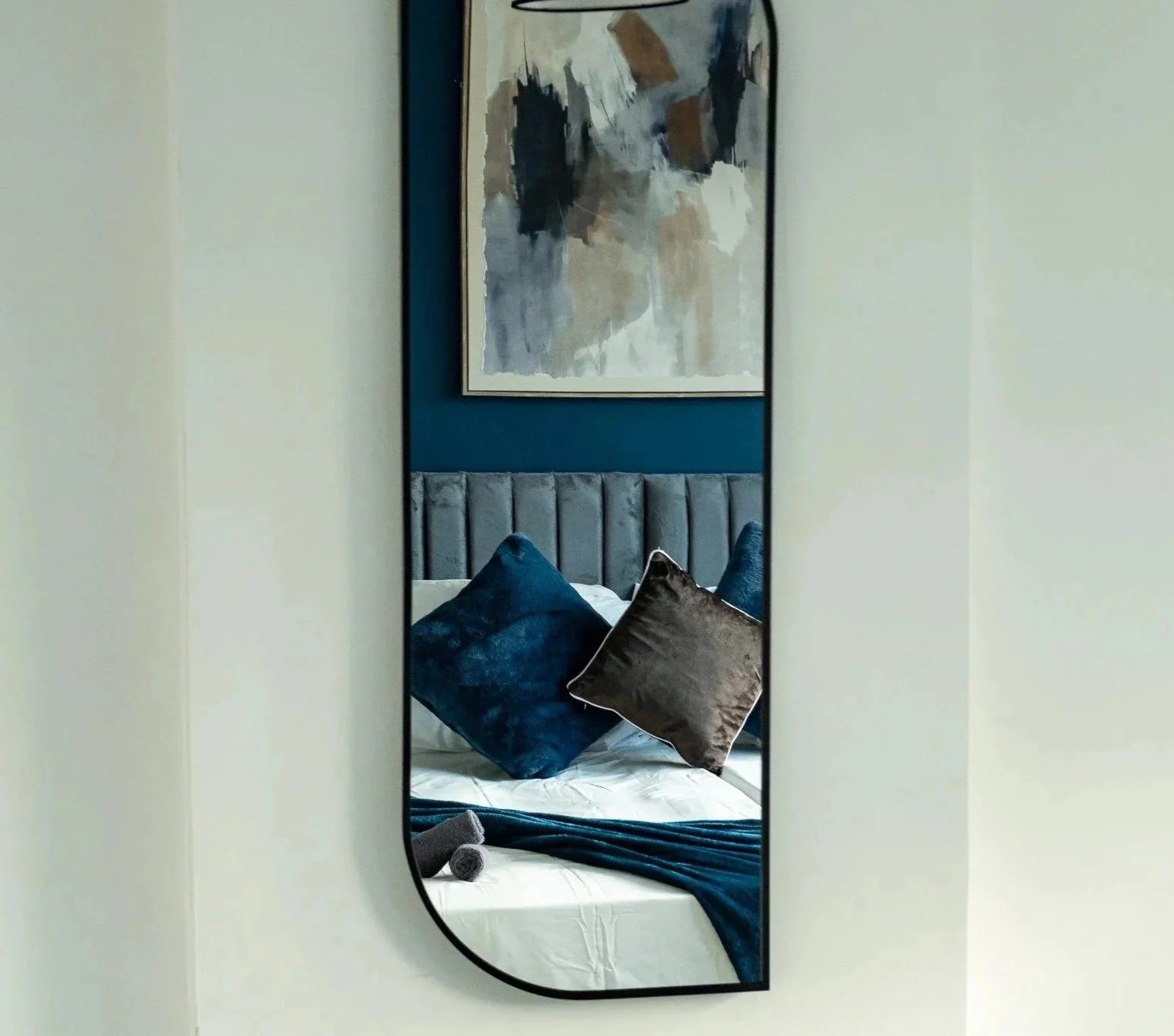 Reflection of a bed with multiple pillows and a headboard, framed in a mirror on a wall with a large abstract painting.