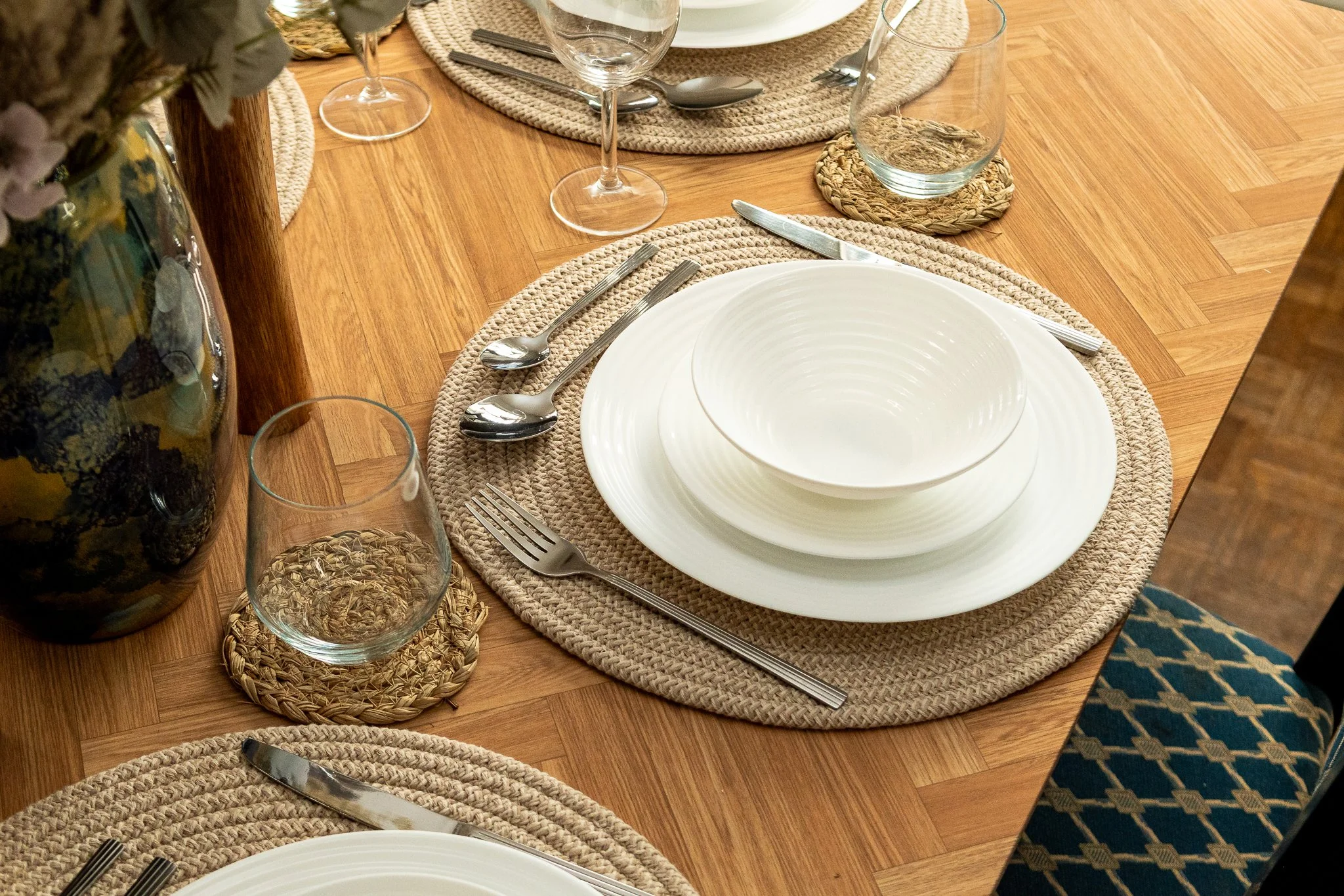 A dining table set with white plates, silverware, clear glasses, and woven placemats on a wooden floor, with a large glass vase on the left containing flowers.