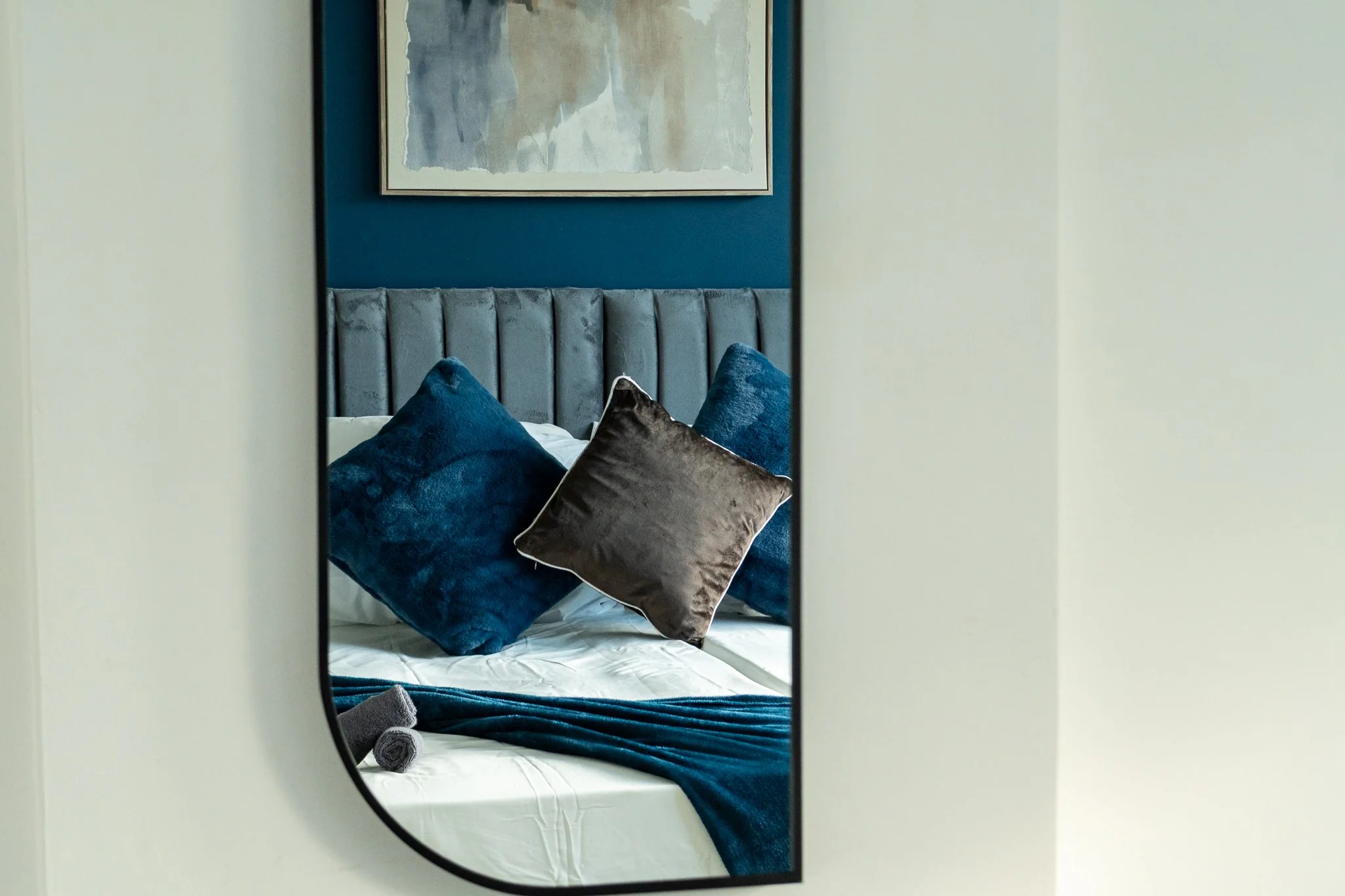 A hotel bed with blue pillows, gray pillows, and a blue bed runner, reflected in a mirror mounted on a wall with a large abstract art piece above the headboard.