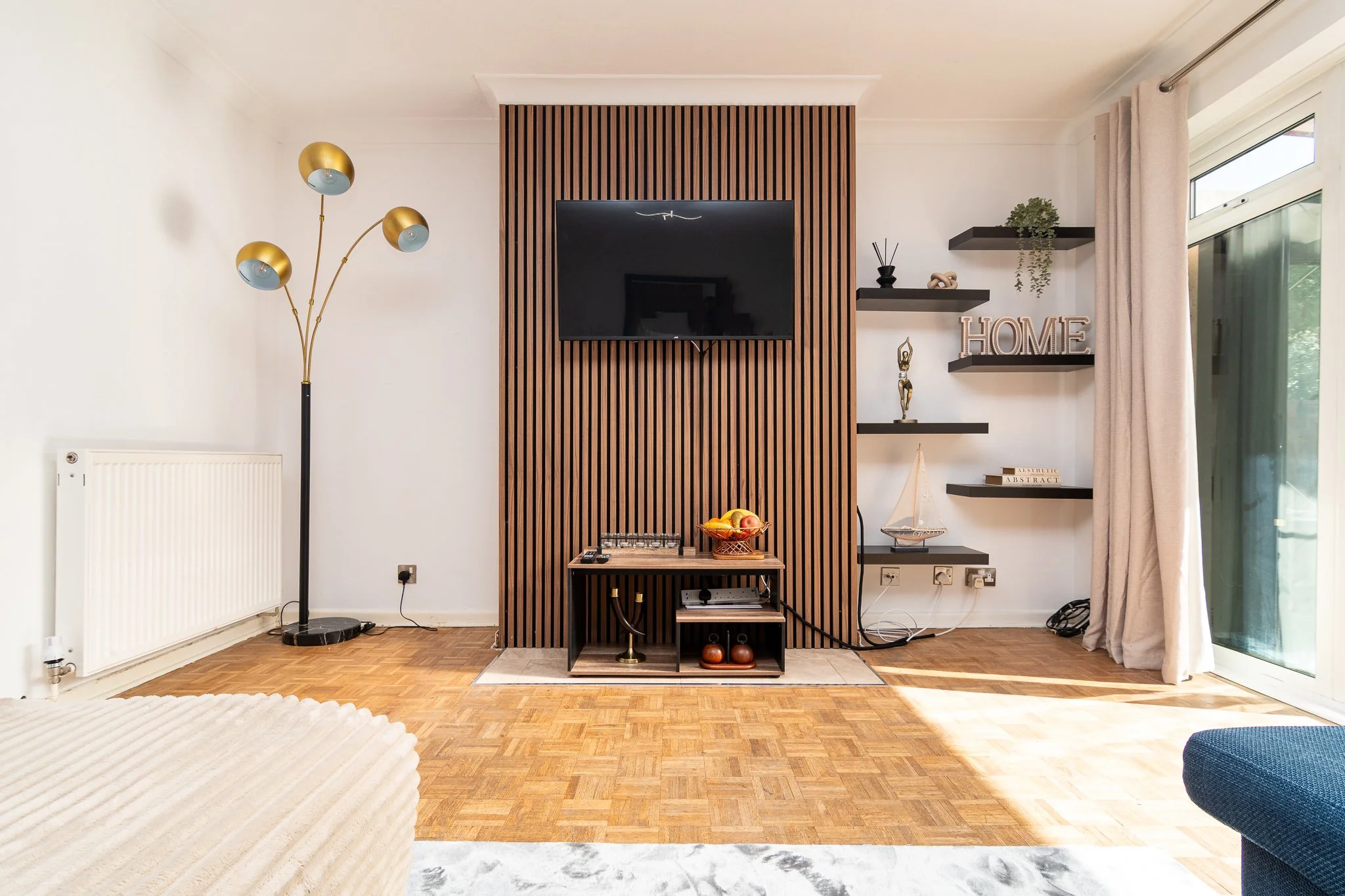 Living room with white walls, wooden parquet floor, and a modern wooden slat TV wall. A black flat-screen TV is mounted on the wall. To the right, floating black shelves hold decorative items and the word 'HOME'. To the left, a multi-arm floor lamp with gold and blue shades. Part of a cream-colored sofa with a rounded armrest is visible in the foreground, and a large window with beige curtains is on the right, providing natural light.
