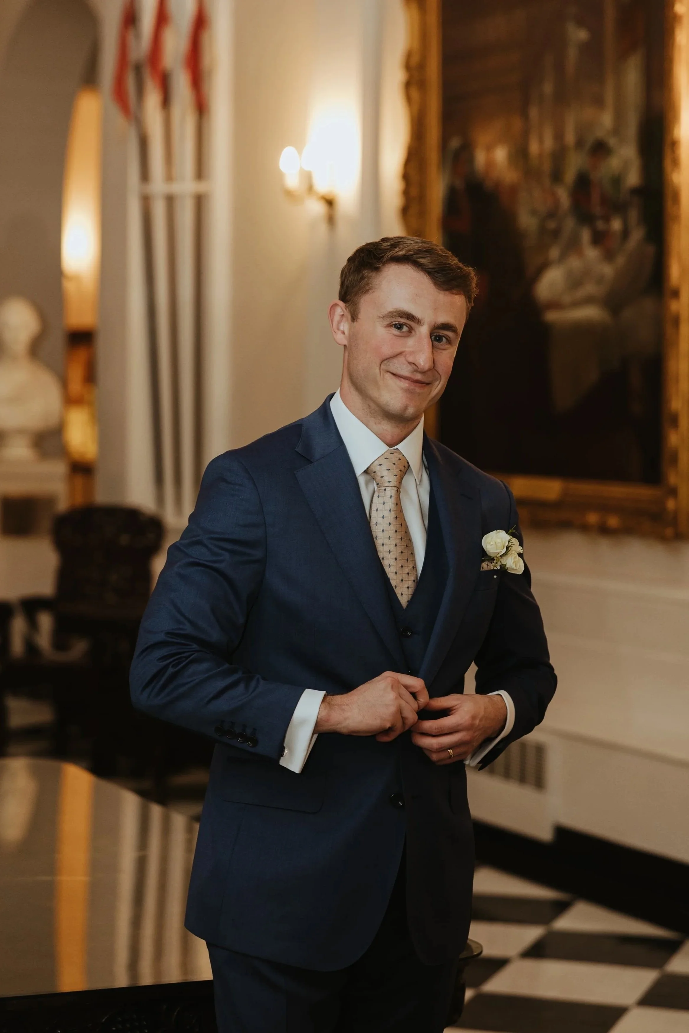 A young man in a blue suit with a white shirt and a beige patterned tie, standing inside an elegant room with ornate decor, a large framed painting behind him, and American flags on the left. He is smiling and holding his hands together.