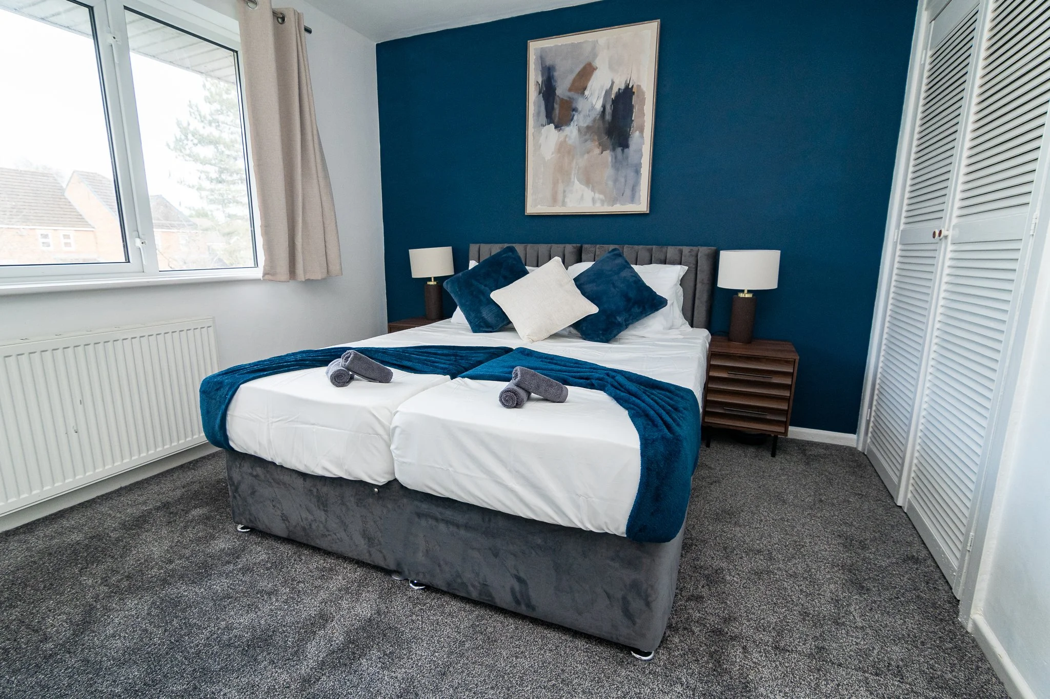 A neatly made hotel room bed with white sheets, a blue velvet blanket, and multiple pillows in shades of blue and white, with two rolled towels on top. There are two wooden nightstands with lamps on either side of the bed, a large abstract painting on the wall behind it, a window with beige curtains, and a white closet with louvered doors.