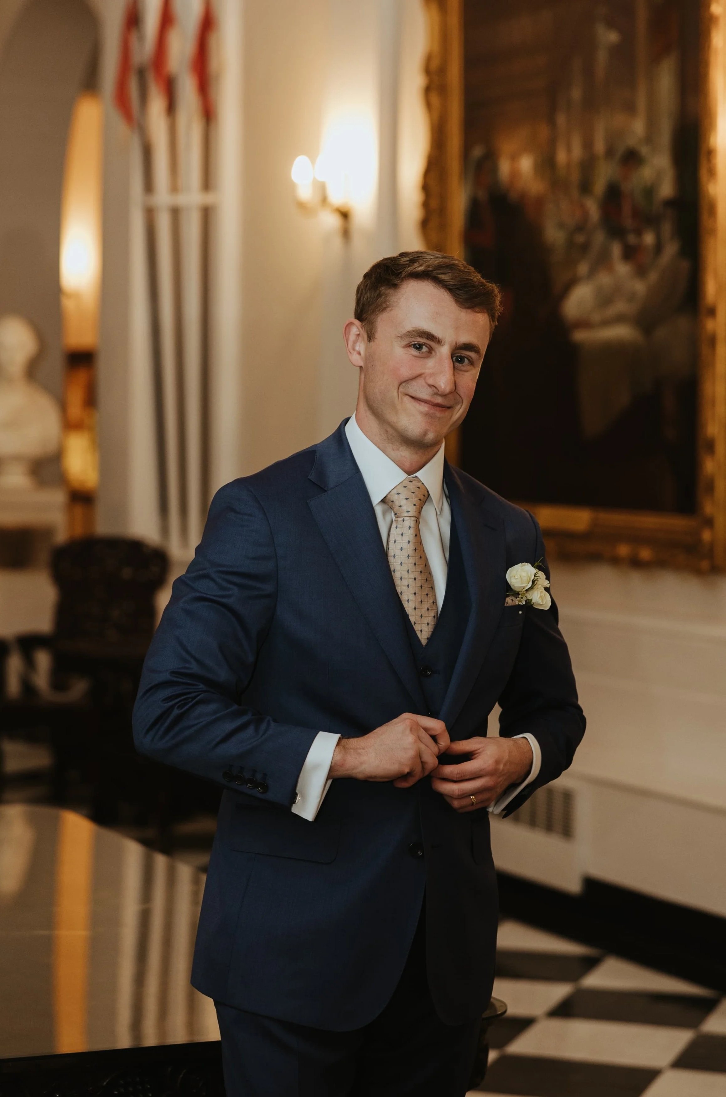 A young man in a dark blue suit with a patterned beige tie and white shirt, wearing a white boutonniere, standing in a luxurious room with gold gilded mirrors, artworks, and classical decor, smiling at the camera.