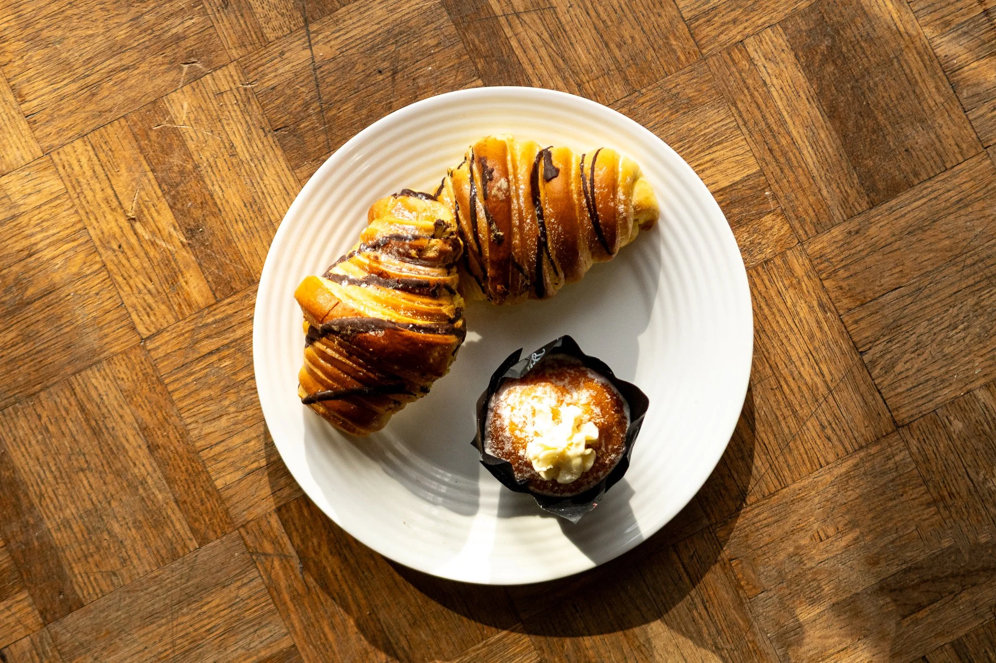 A white plate with three baked goods and a small cupcake on a wooden table. The baked goods are topped with chocolate drizzle and sugar.