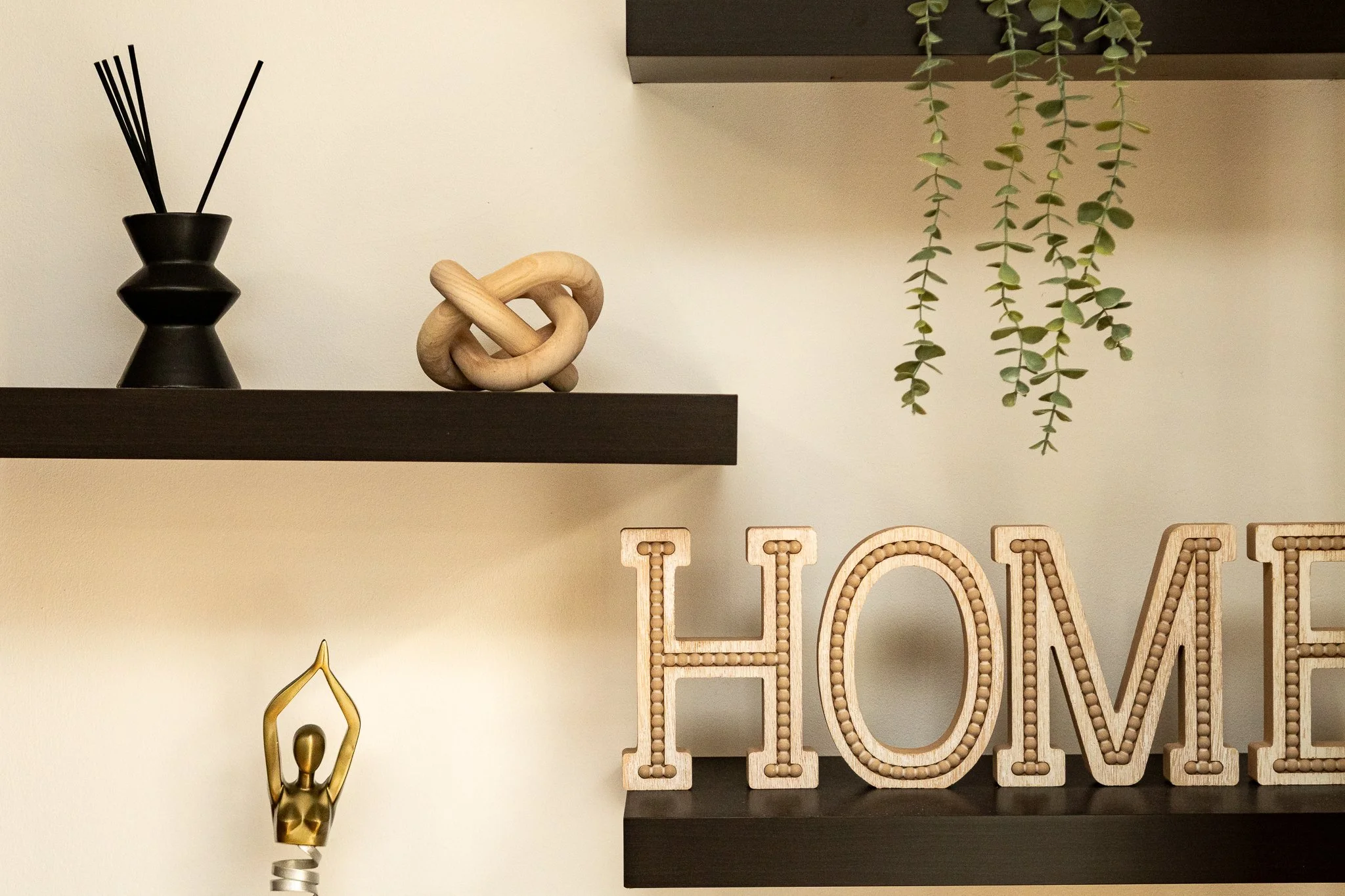 Decorative wall shelf with black vase holding reed diffuser sticks, a wooden knot sculpture, hanging green trailing plants, a gold deer head figurine, and large wooden letters spelling 'LOVE'