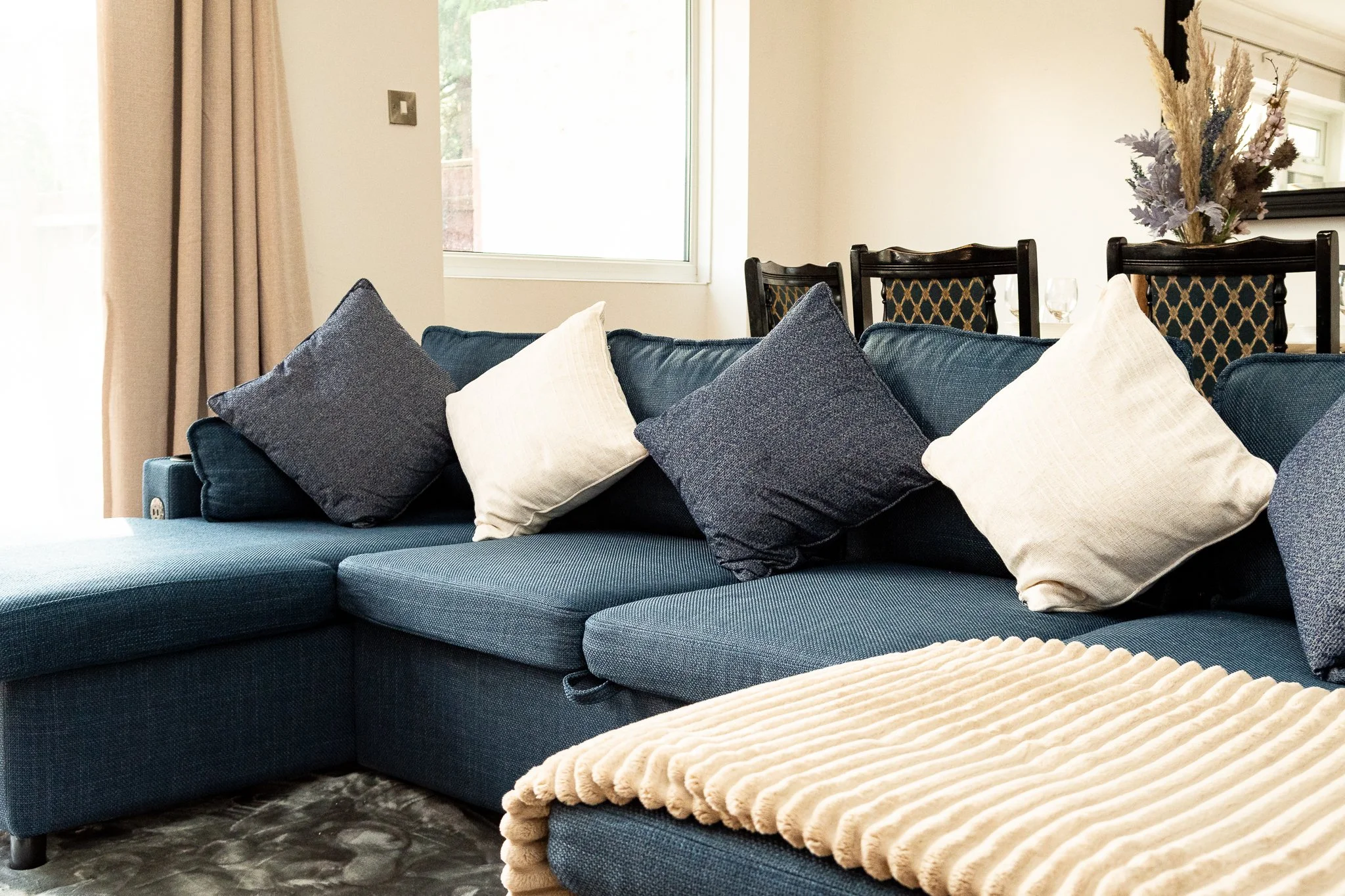 A dark blue sectional sofa with multiple blue and white pillows, a beige textured blanket on the ottoman, a window with beige curtains, and a dining area with chairs and a floral arrangement in the background.