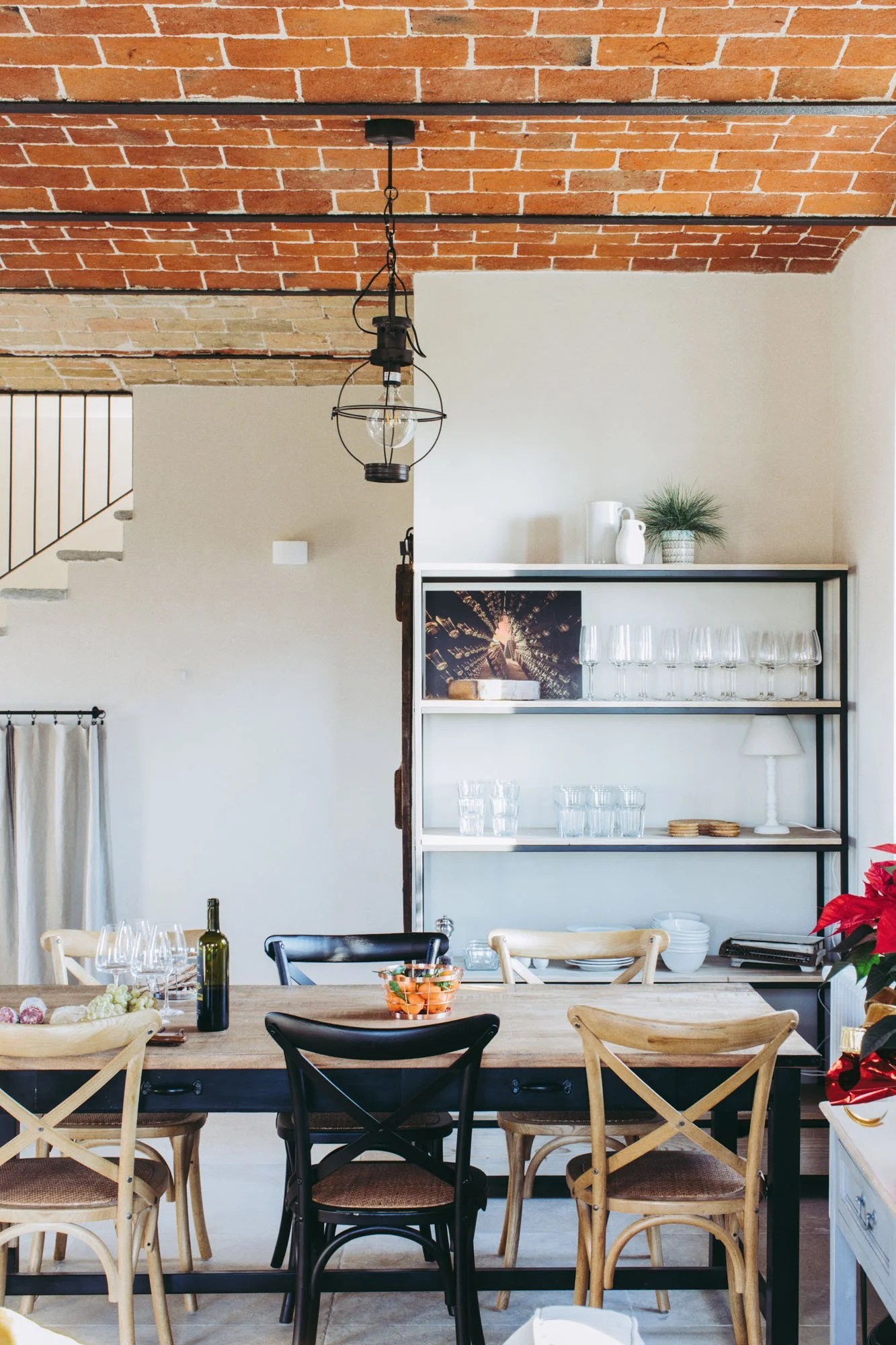 Interior of a dining room with a wooden table set with wine glasses, a wine bottle, and a bowl of oranges. The background features a white wall with a shelving unit holding glassware, vases, and a plant, and a brick ceiling with a hanging light fixtu
