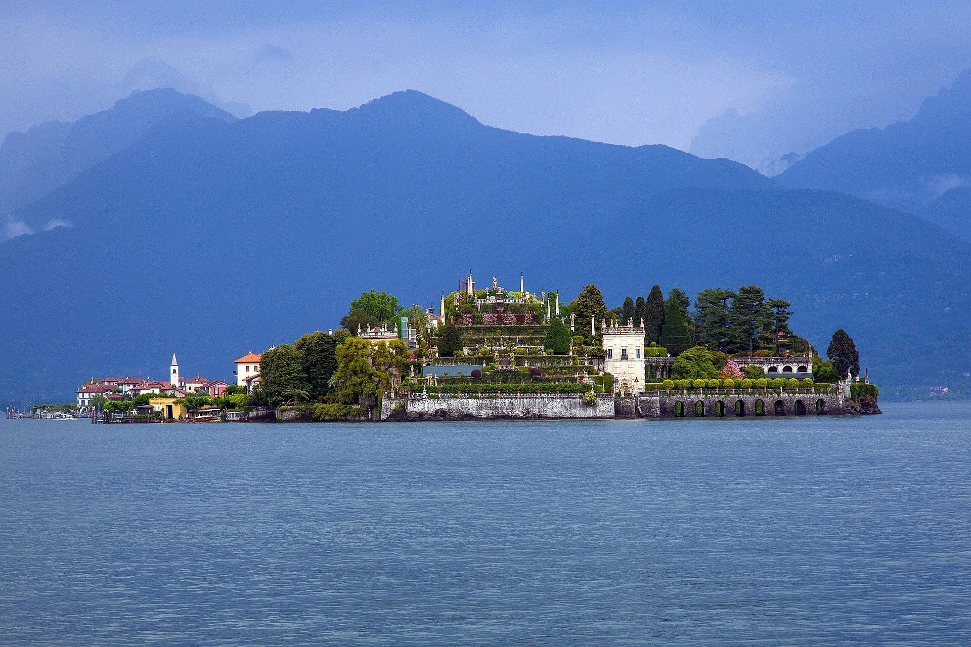A large historic villa-like building on an island in lake Maggiore, surrounded by water, with a backdrop of mountain range and cloudy sky.
