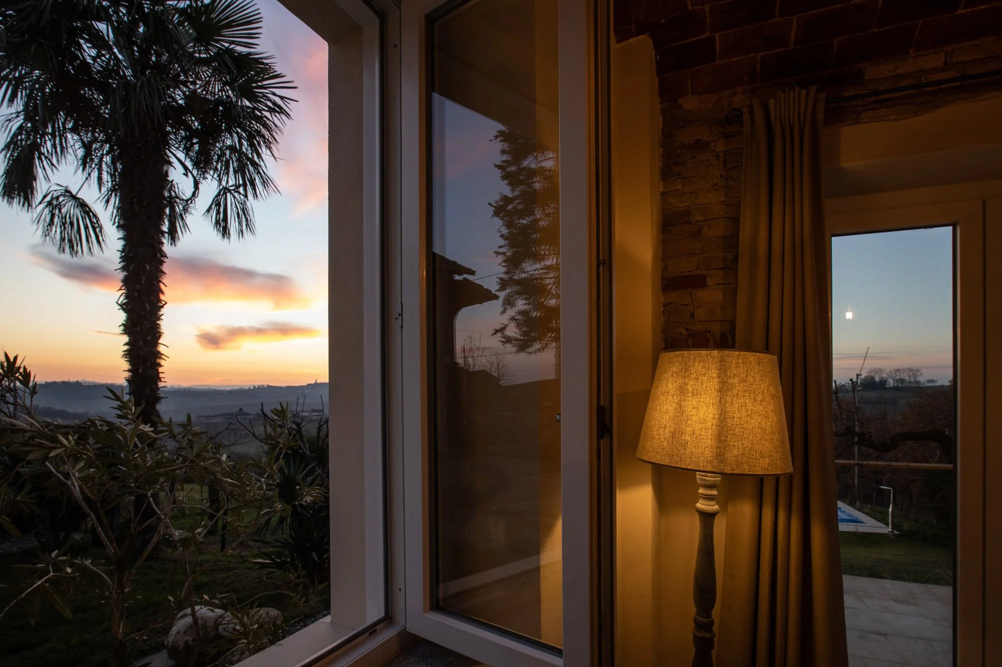 Interior view of a room with a floor lamp, curtains, and large windows revealing trees and a sunset sky outside.