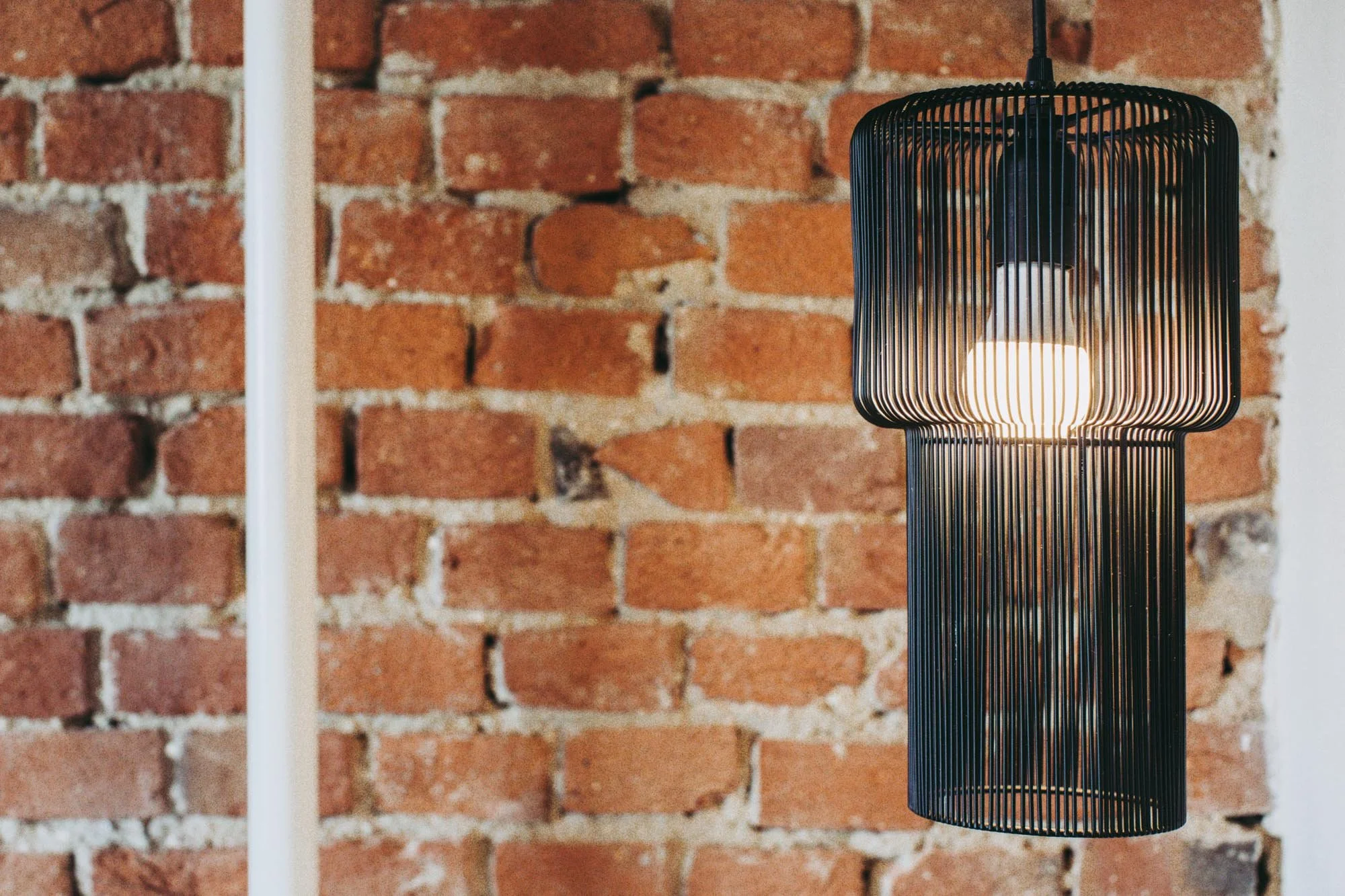 A black wire pendant light fixture hanging in front of a red brick wall.