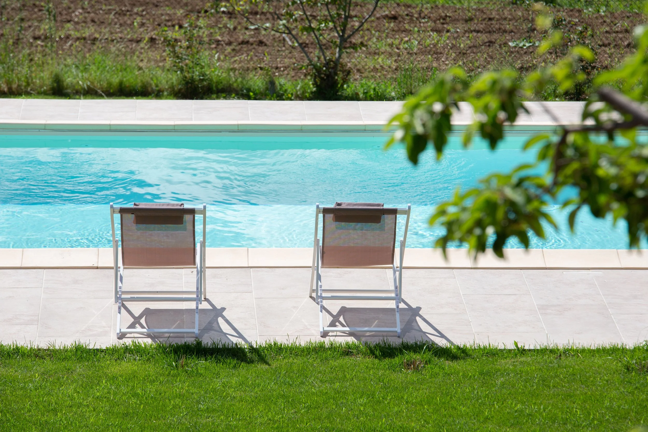 Two lounge chairs by a swimming pool overlooking a backyard with trees and grass.