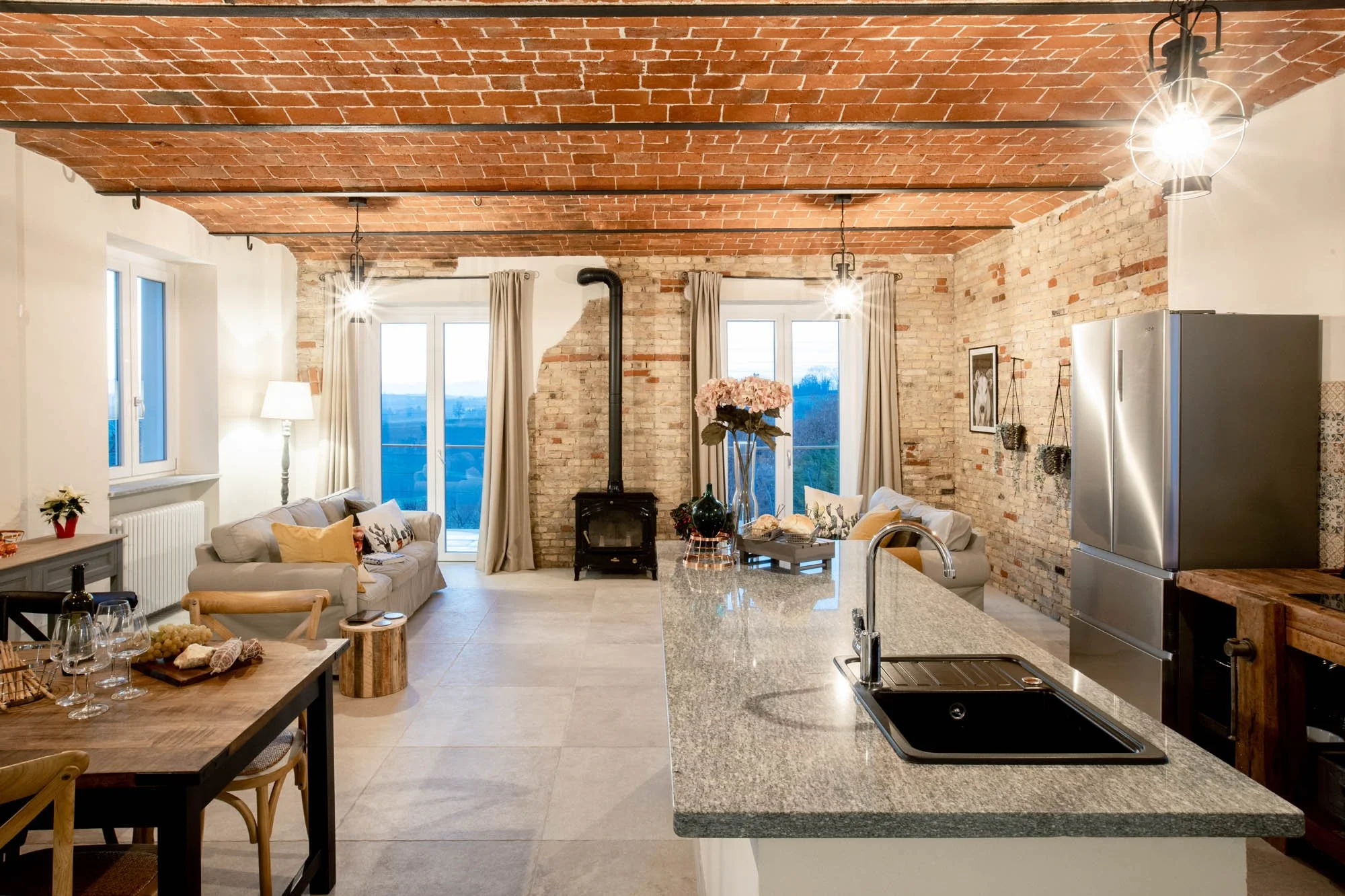 A cozy living room with brick walls and ceiling, large windows with beige curtains, a white sofa with patterned cushions, a wood stove, and a wooden dining table with wine glasses on it. Kitchen with granite countertop and stainless steel refrigerato