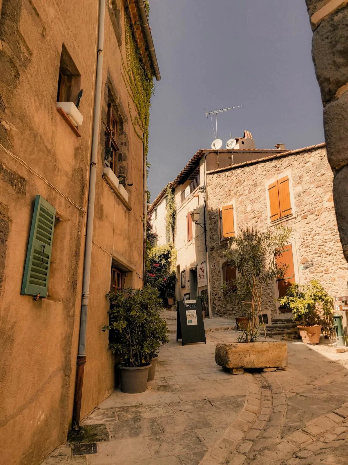 A narrow, cobblestone street in an Italian village with stone and stucco buildings, some with shuttered windows and small potted plants outside. The sky is cloudy in the background.