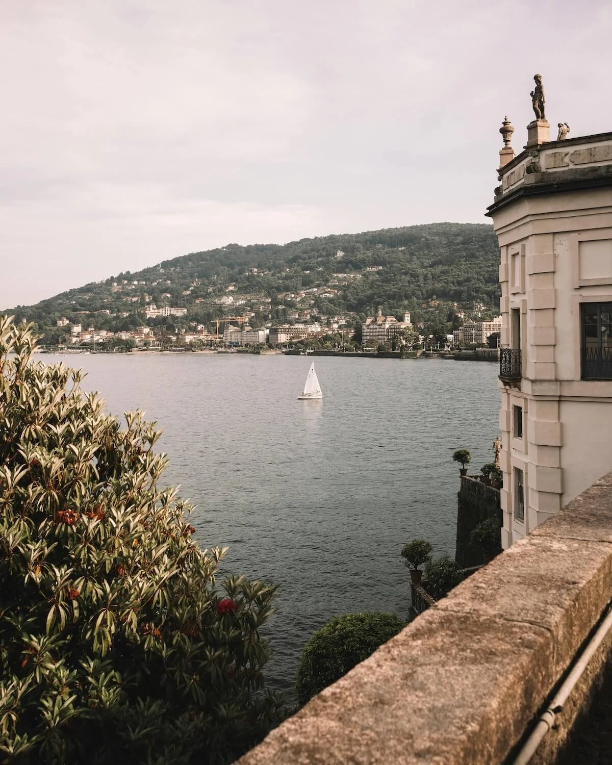 A scenic view of a river with a sailboat in the middle, surrounded by a hillside with numerous buildings. On the right side, part of a white historic building with statues on the roof is visible. In the foreground, green leafy bushes and a stone railing are present.
