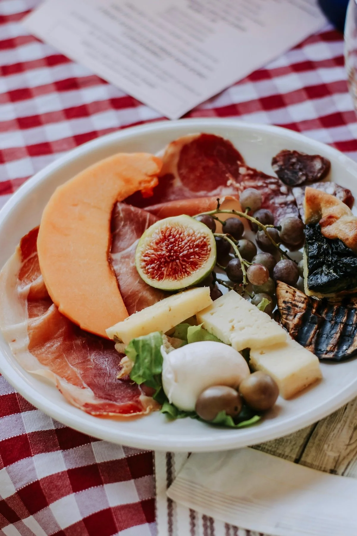 A plate of assorted cheeses, cured meats, fresh grapes, and a halved fig on a red and white checkered tablecloth.