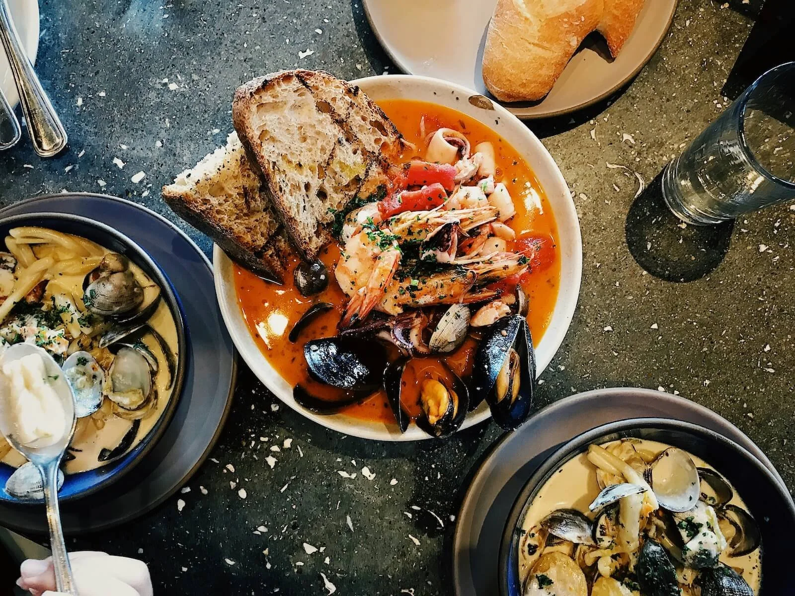 Seafood dishes including a bowl of seafood stew with bread, clams and mussels in a creamy sauce, and another bowl of seafood pasta with clams and mussels, on a black speckled table.