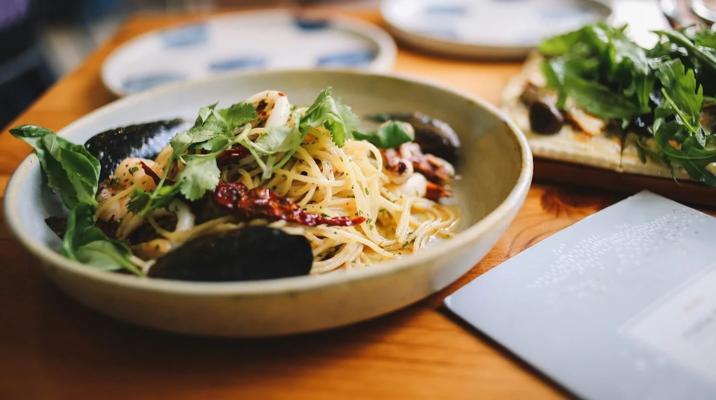 A bowl of pasta with black mussels, cherry tomatoes, and fresh greens on top, placed on a wooden table with other dishes in the background.