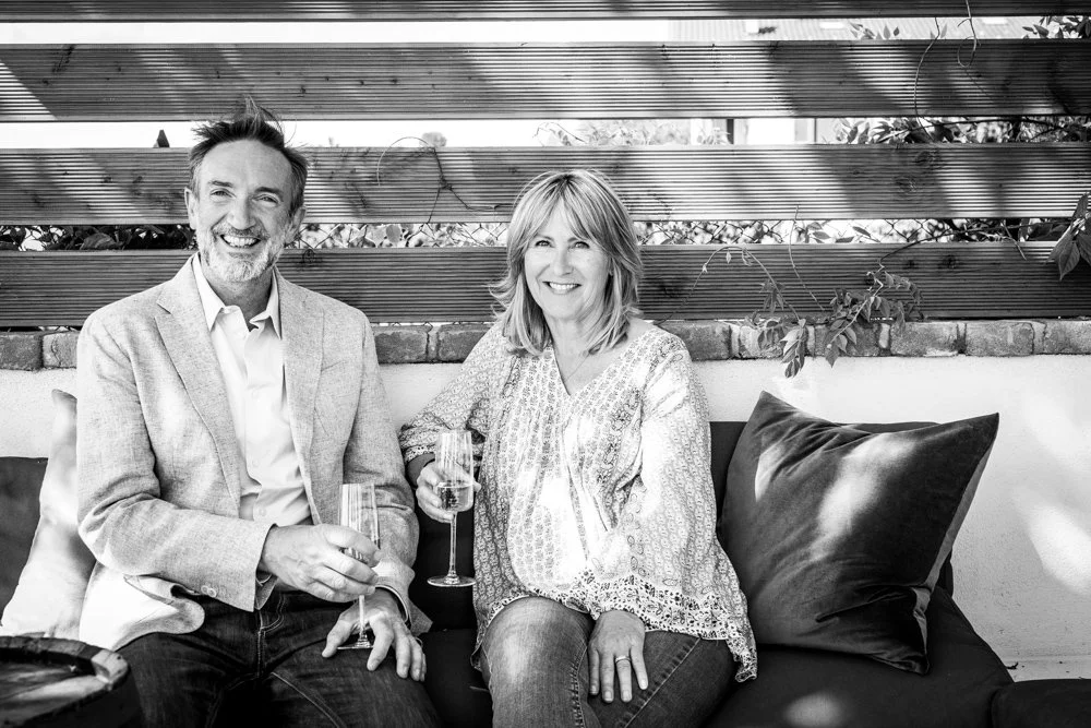 A smiling man and woman sitting on a sofa with glasses of champagne, in an outdoor setting with plants and wooden slats behind them.