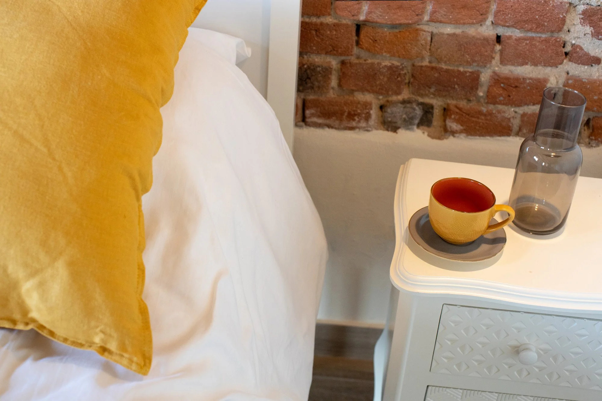 A yellow mug, a clear glass bottle, and a small dark tray are placed on a white bedside table next to a bed with white sheets and a yellow pillow. A brick wall is visible in the background.