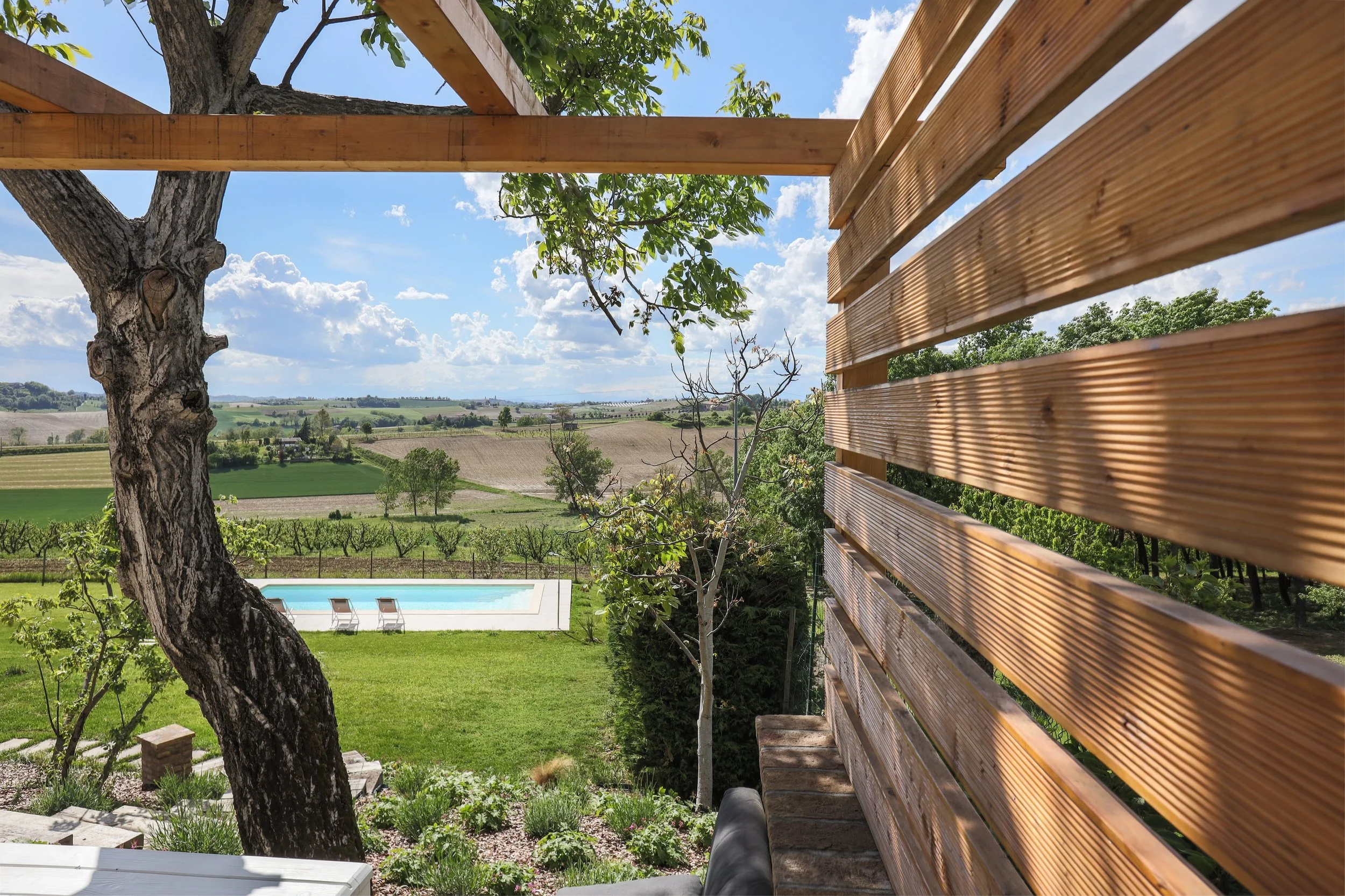 View from a balcony showing a swimming pool and lawn, with trees, fields, and a blue sky with clouds in the background, and a wooden privacy screen on the right side.