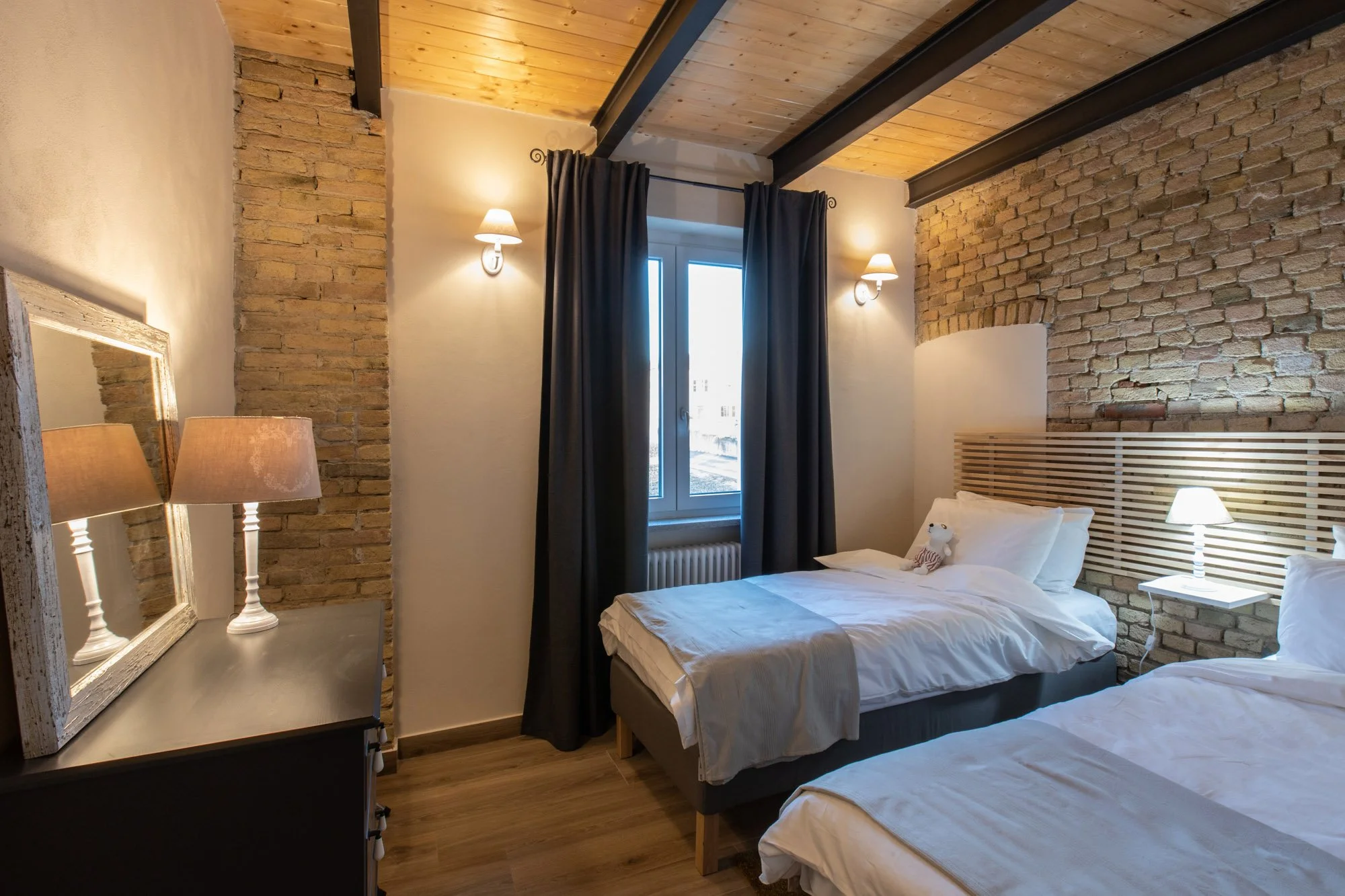 A bedroom with brick and white walls, wooden ceiling, black beams, and dark curtains. There are two beds with white bedding, bedside lamps, and a window with sunlight streaming in.