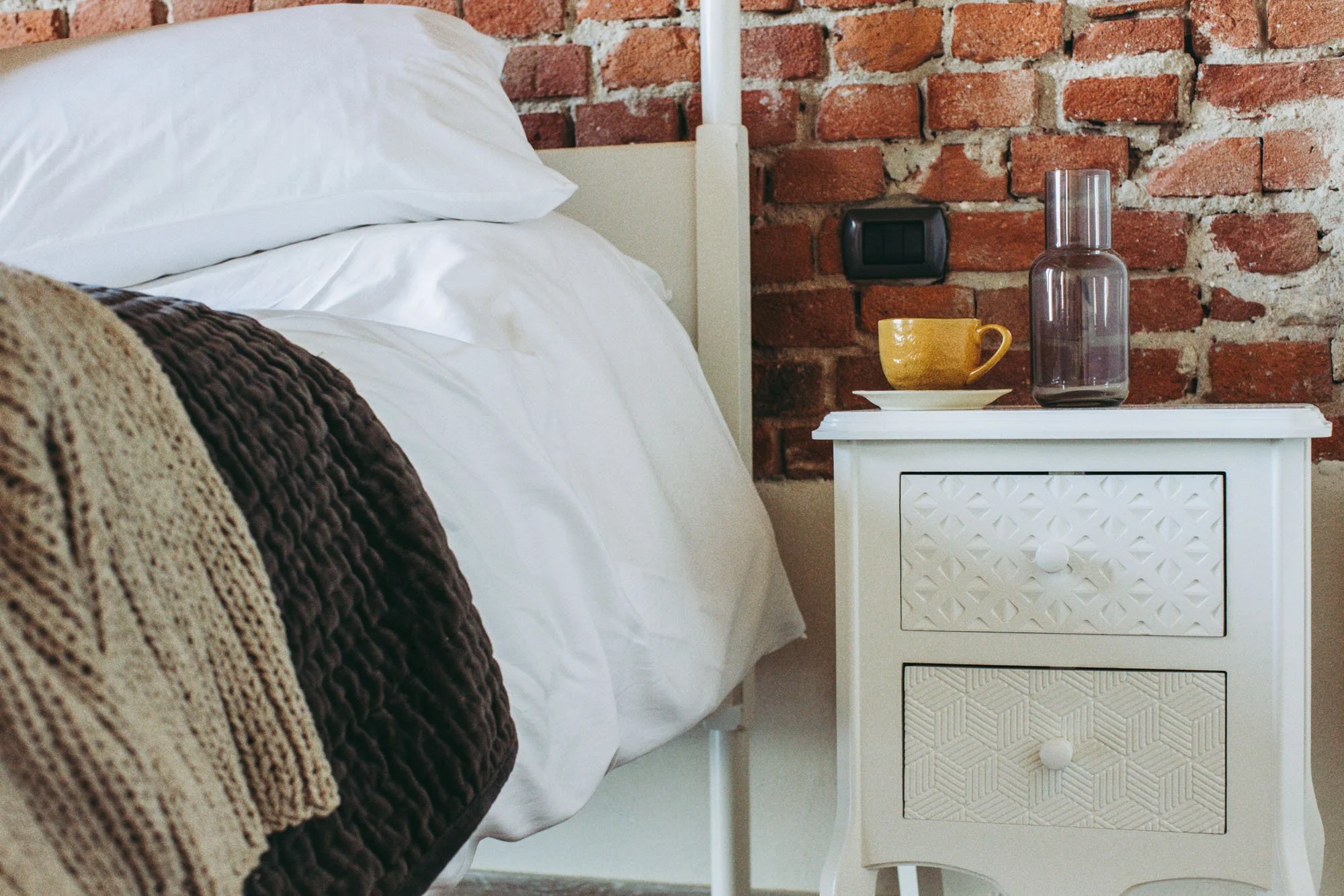 Part of a cozy bedroom with white bedding, a knitted brown blanket, and a cream-colored nightstand with a yellow cup, saucer, and a purple glass vase against a red brick wall.