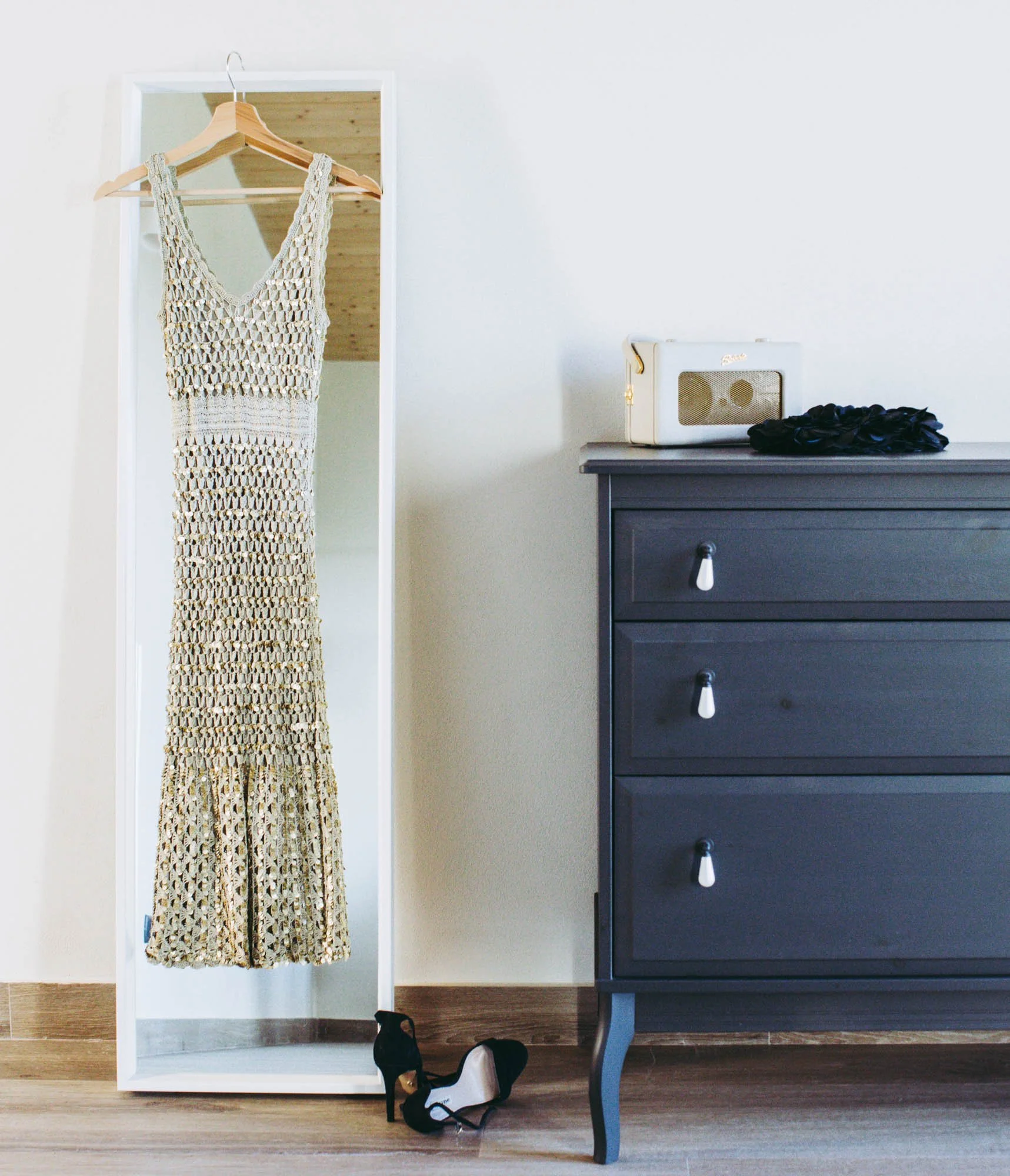 A boho style crochet dress hanging on a wooden hanger in front of a mirror, with black and white shoes and a black and white bag on the floor, and a black dresser on the right side.