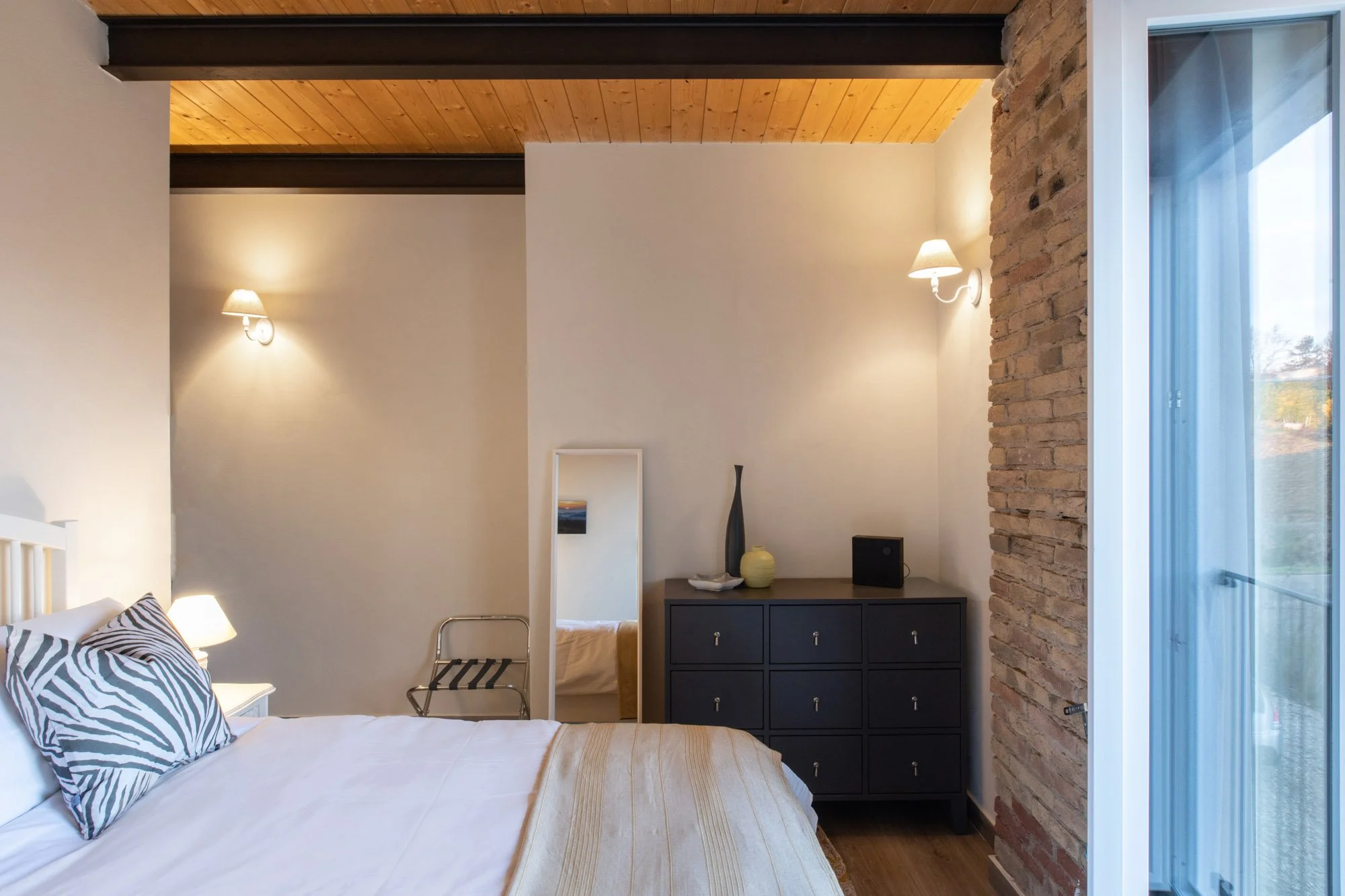 A bedroom with a wooden ceiling and brick wall, featuring wall-mounted lamps, a bed with zebra-print pillows, a small mirror, and a black dresser with decorative items. A sliding glass door is visible on the right, revealing an outdoor view.