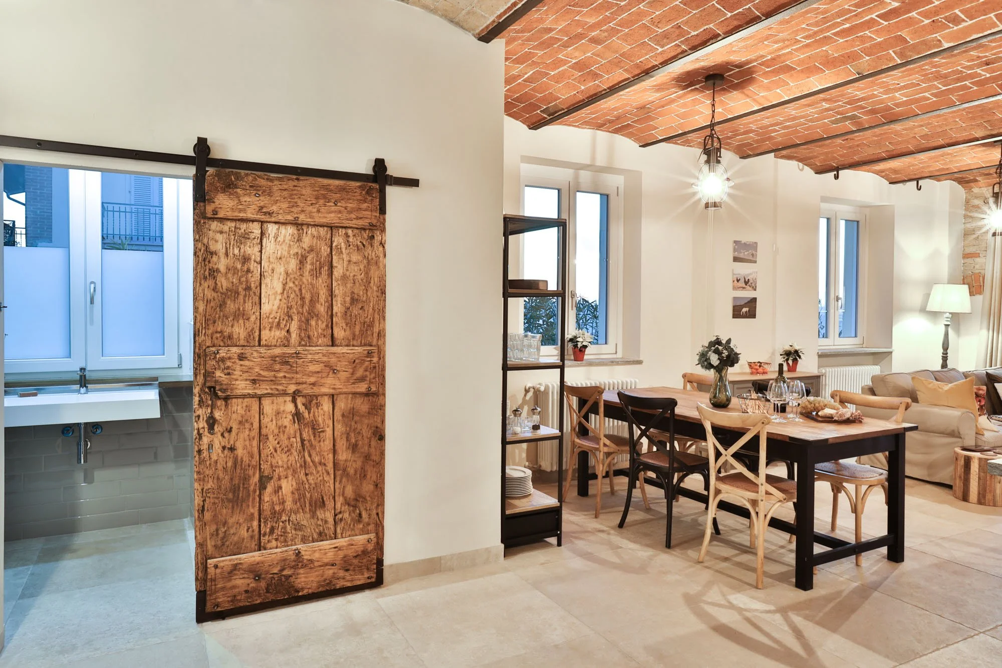 Interior of a dining room with a brick vaulted ceiling, white walls, and large windows. Visible are a wooden dining table with chairs, a table lamp, and some wall decorations. A sliding barn door separates a kitchen area with a sink and two windows.
