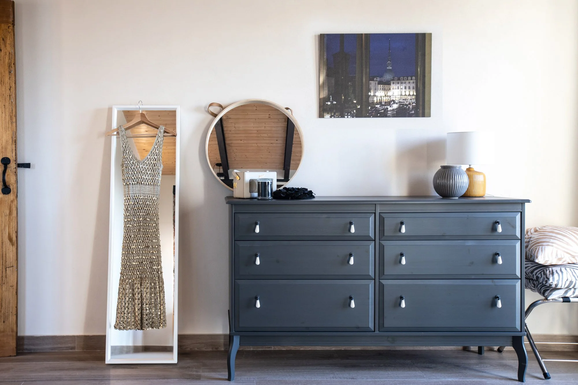 A bedroom with a dark blue dresser, a round mirror, and a decorative lamp. A dress with a crochet pattern is hanging on a white full-length mirror. A framed cityscape photograph is on the wall.