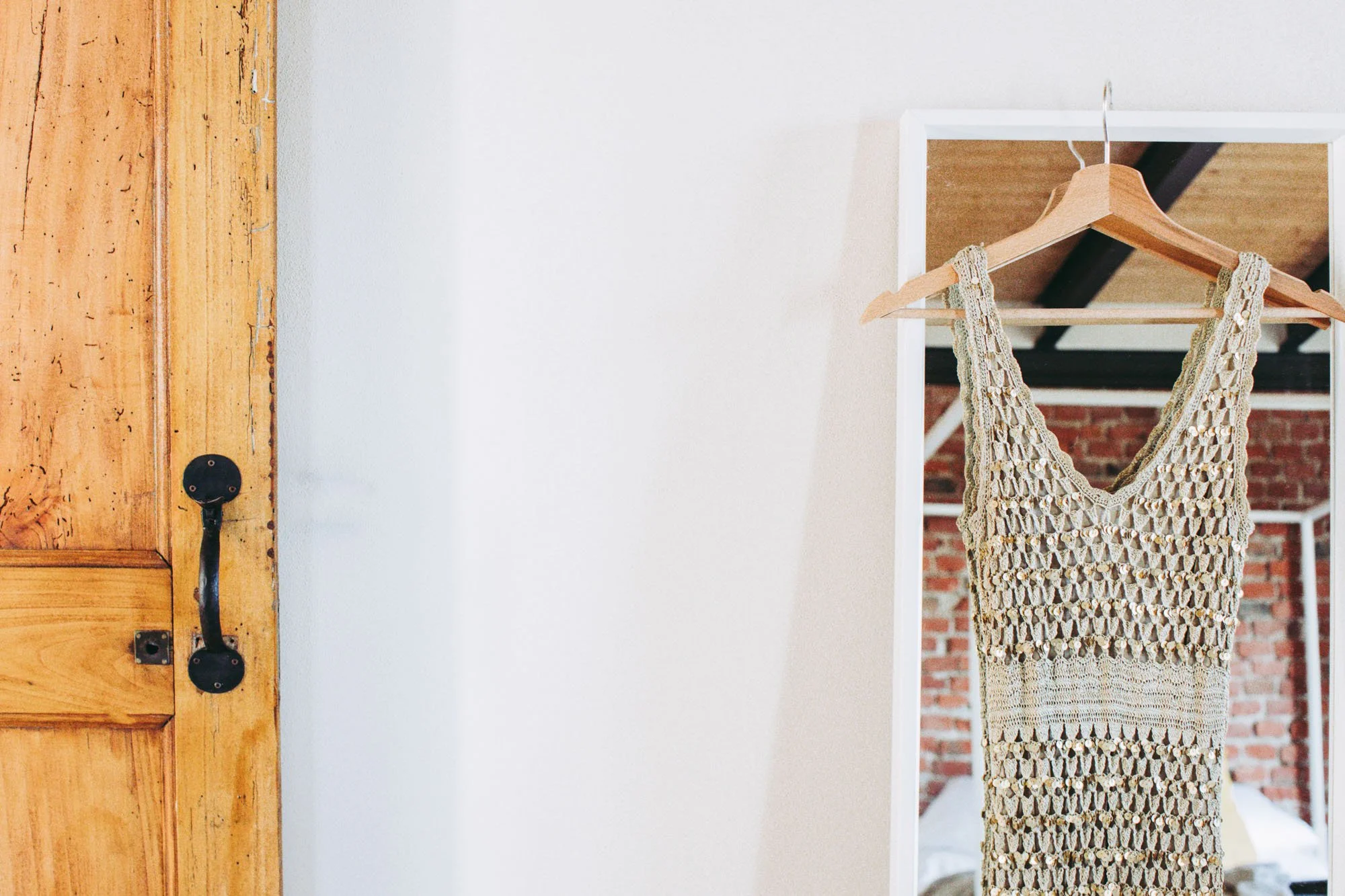 A beige crocheted dress on a wooden hanger hanging on a hook inside a white-framed mirror, with a brick wall in the background.