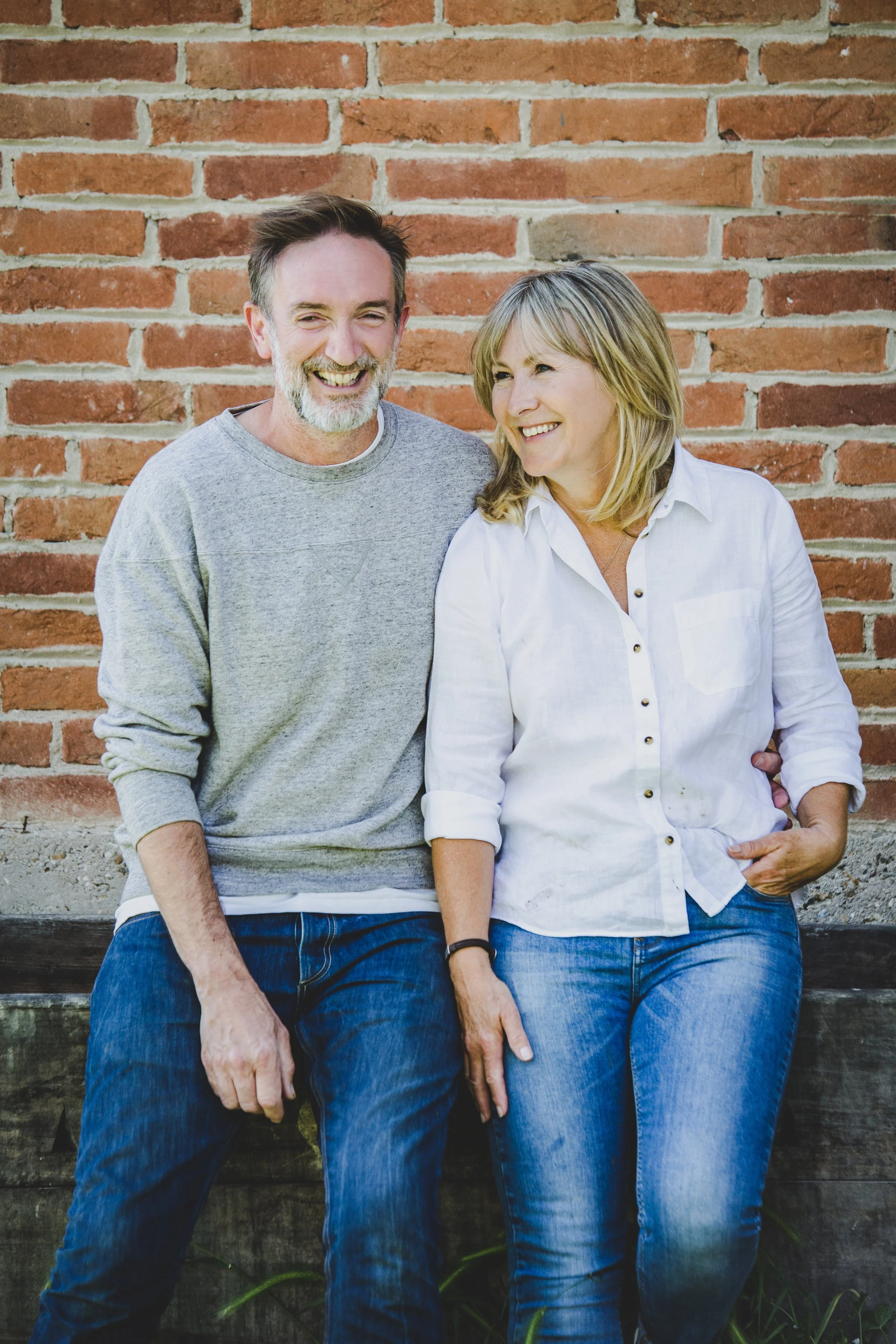 A smiling middle-aged man with a beard and a woman with shoulder-length blonde hair sitting against a brick wall, wearing casual clothing.