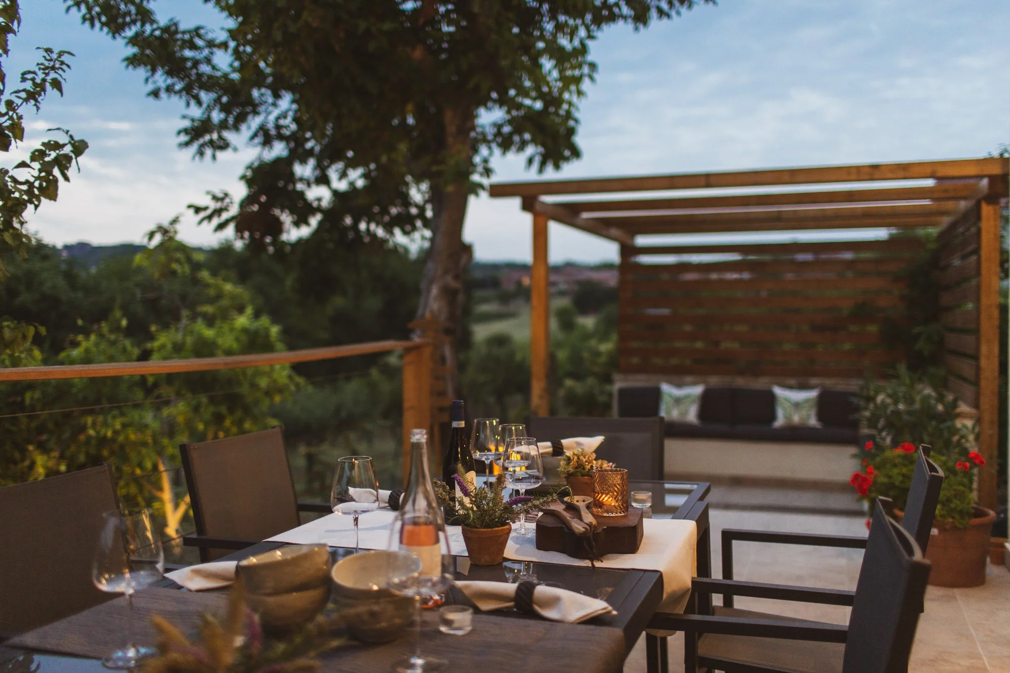 Outdoor dining area at dusk with a table set for a meal, including wine glasses, plates, and a candle, on a deck with scenic view of trees and a cloudy sky.