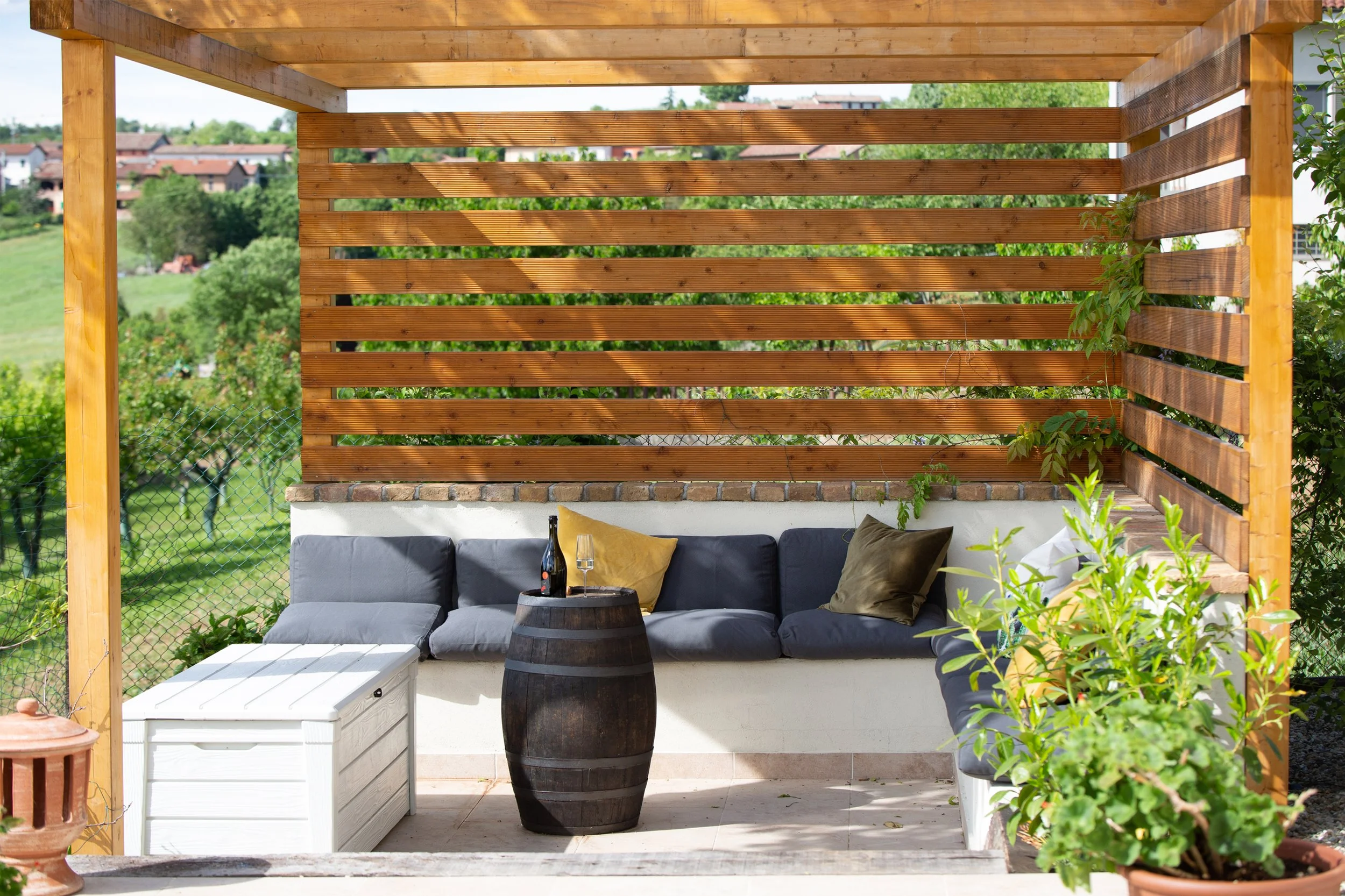 Outdoor patio with a wooden pergola, gray sofa with pillows, a barrel table with drinks, and potted plants, overlooking a green landscape with houses.