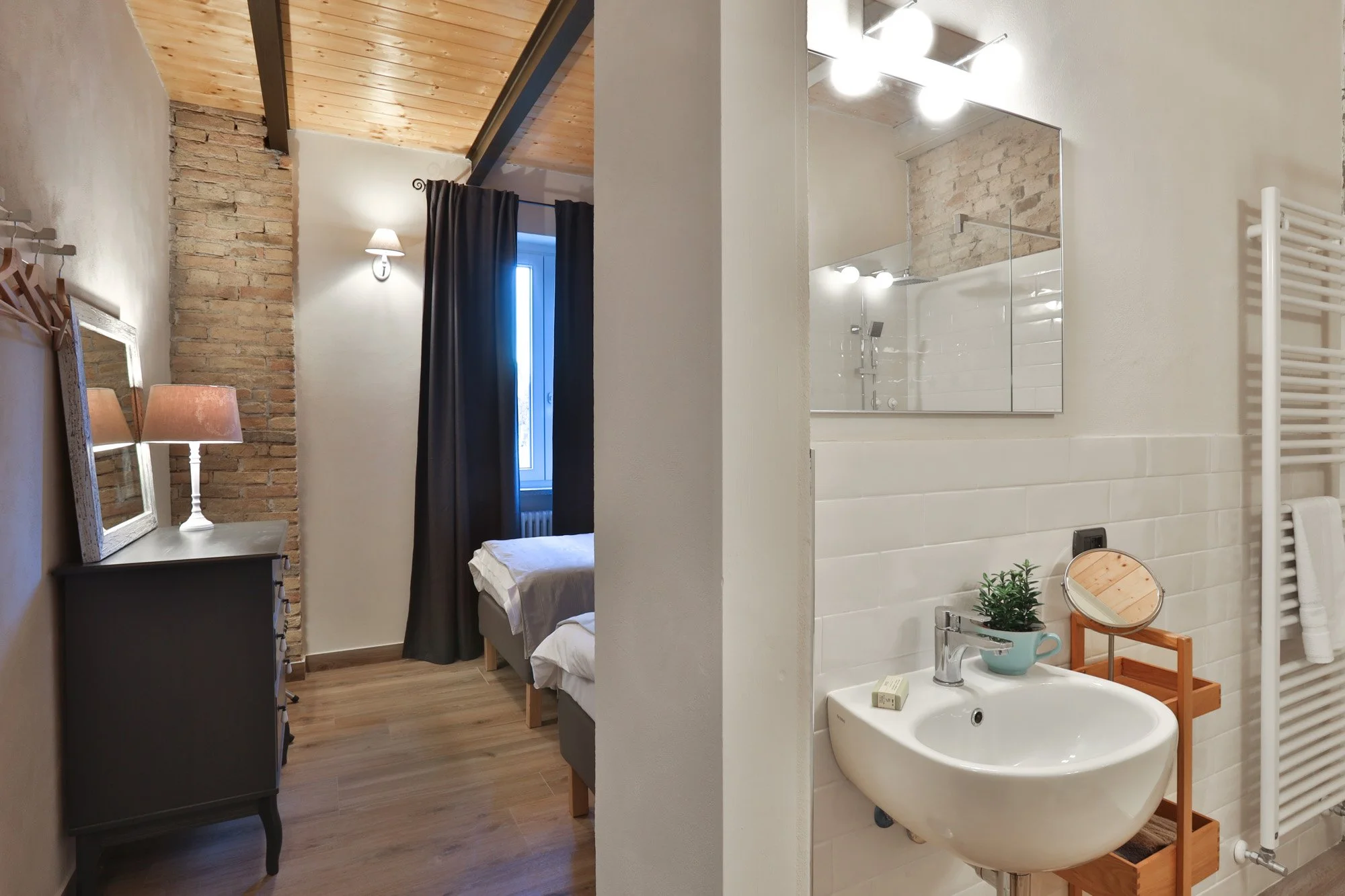 A room split into two sections: the left side shows a bedroom with exposed brick and wood ceiling, a small dark dresser, a table lamp, and a bed, while the right side features a bathroom with a sink, mirror, white tiled wall, potted plant, soap, and 