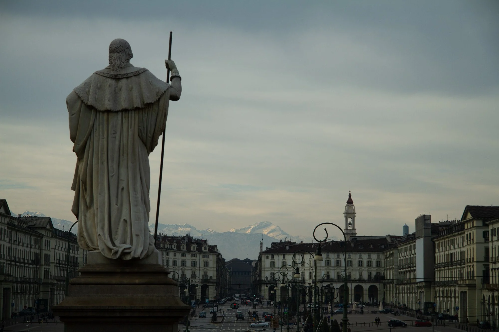 A statue with a staff overlooking Piazza San Carlo in the centre of Turin, which has fabulous architecture & historic buildings, clock towers, and snow-capped mountains in the background under a blue sky.