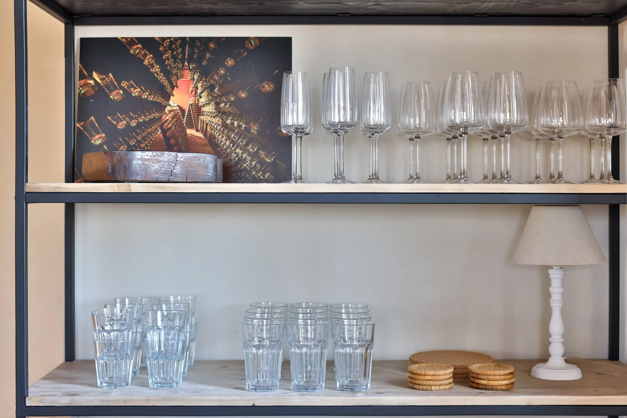 A shelf displaying wine glasses and wooden coasters with a framed picture of a wine cellar in the background, and a small white lamp on the right side of the shelf.