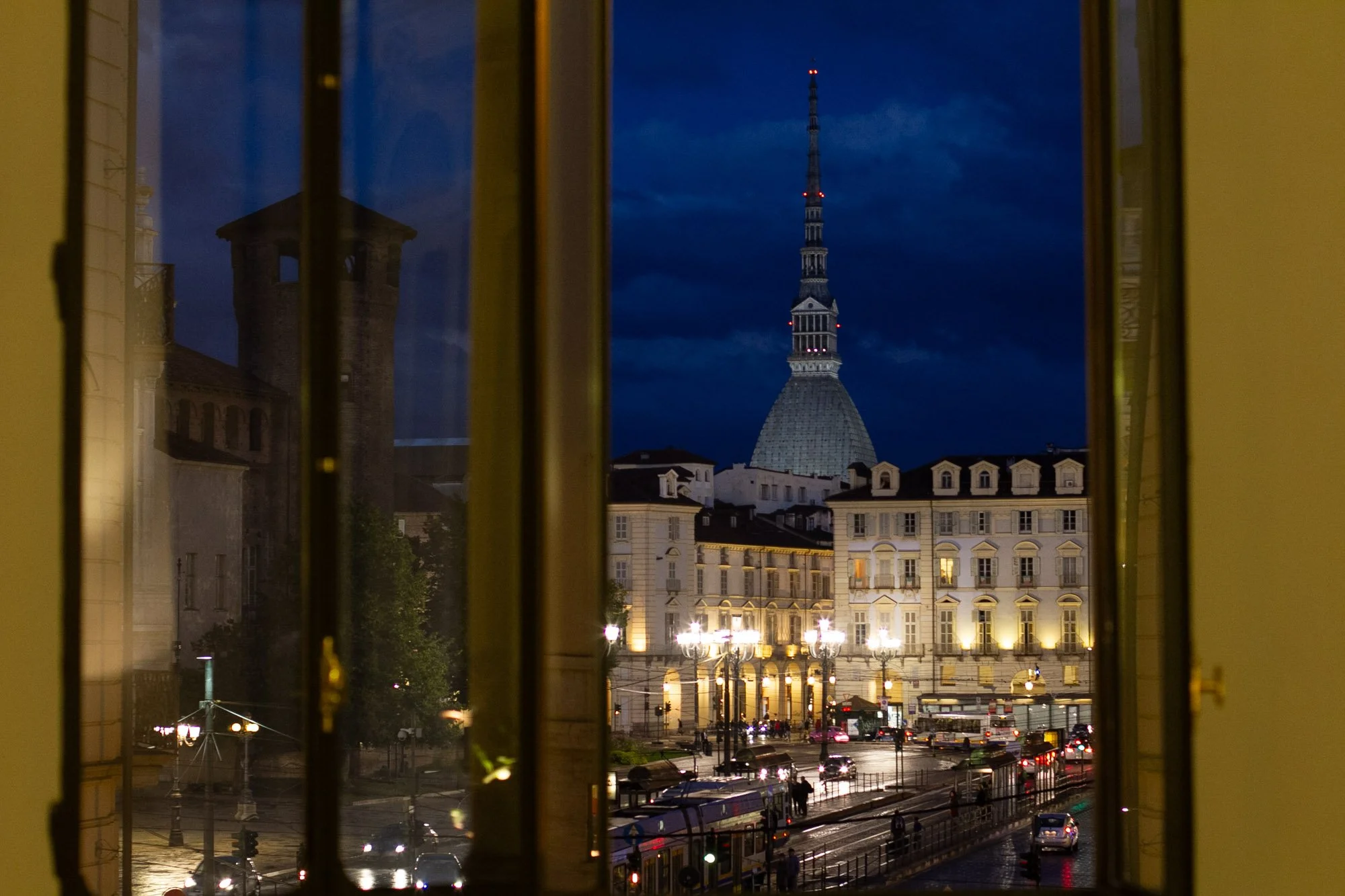 View of the city of Turin, Italy, seen through a window, with the Mole Antonelliana's dome and spire illuminated against a dark blue evening sky.