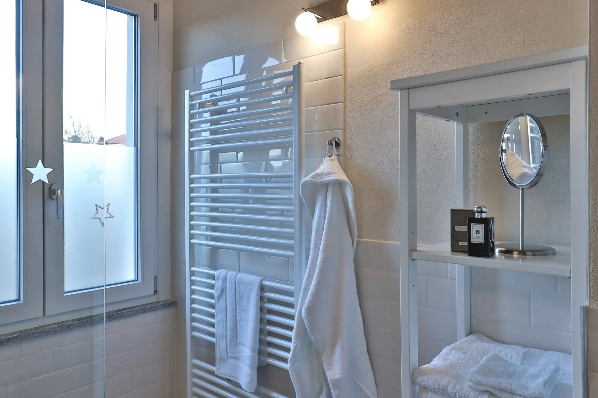 A small, modern bathroom with a glass door with star decals, a white radiator, a towel hanging, a white shelf with perfume bottles, a mirror, and a fluffy white rug.