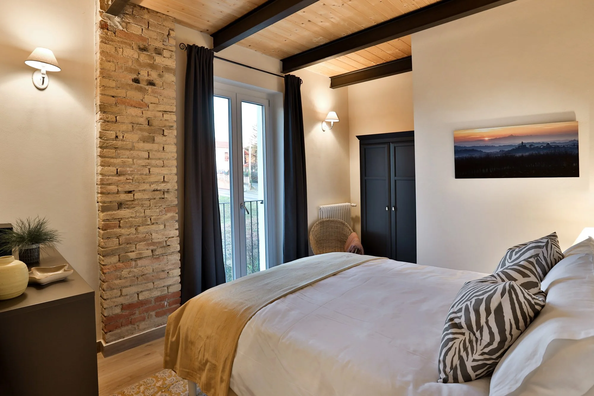 A bedroom with a large bed that has zebra-striped pillows and a beige blanket. There is a dark blue wardrobe, a wicker chair, and a window with black curtains. The room has a rustic ceiling with wooden panels and black beams, brick and white walls, a