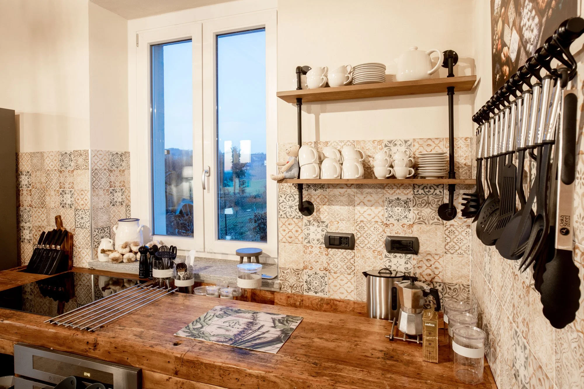 Kitchen with wooden countertop, shelves with coffee mugs, plates, teapot, and kitchen utensils hanging on wall, window showing outdoor view.